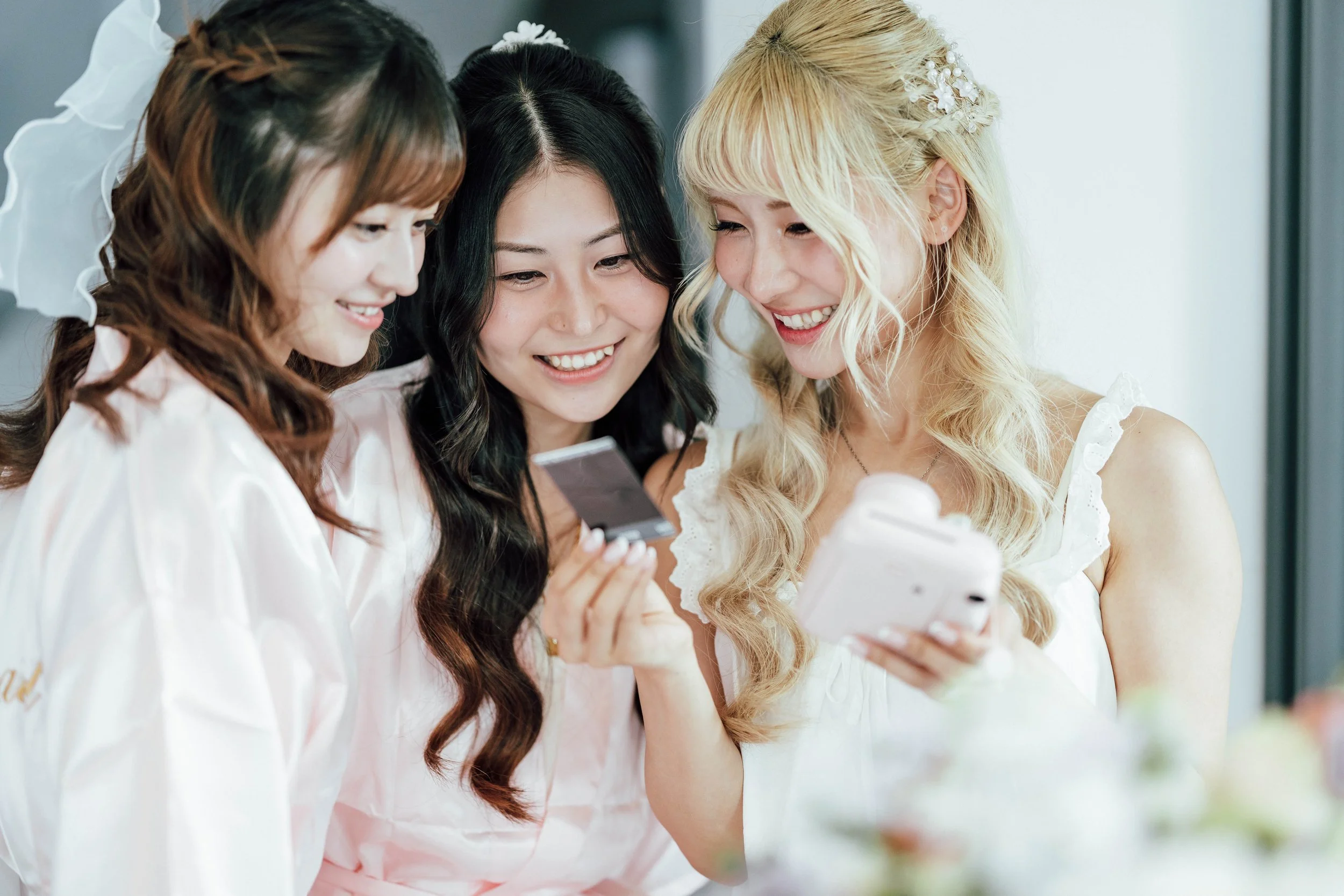 Three women in white dresses looking at a smartphone and smiling, celebrating a wedding.