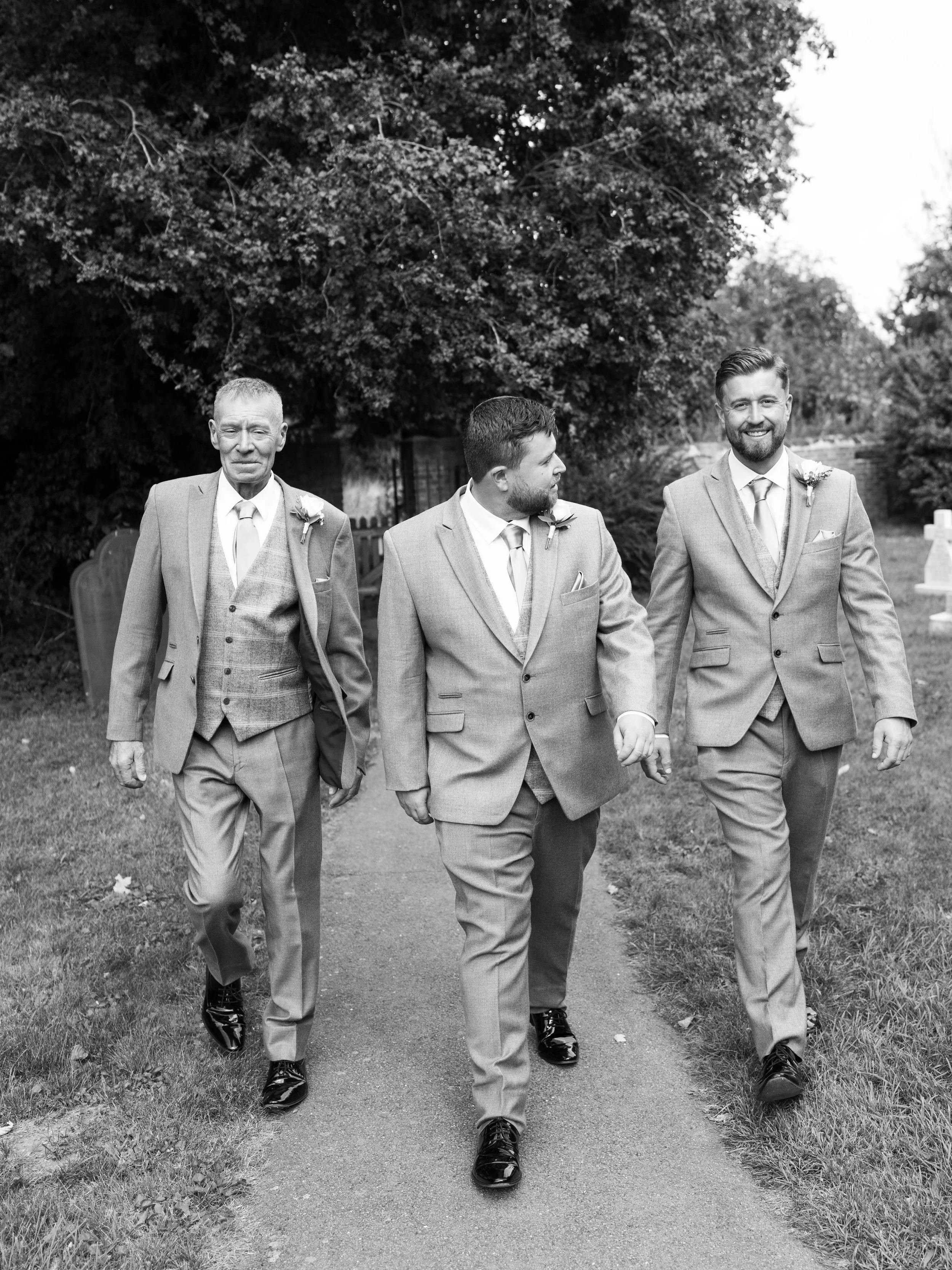 Three men in suits walking outdoors, holding hands, with trees and a bench in the background.