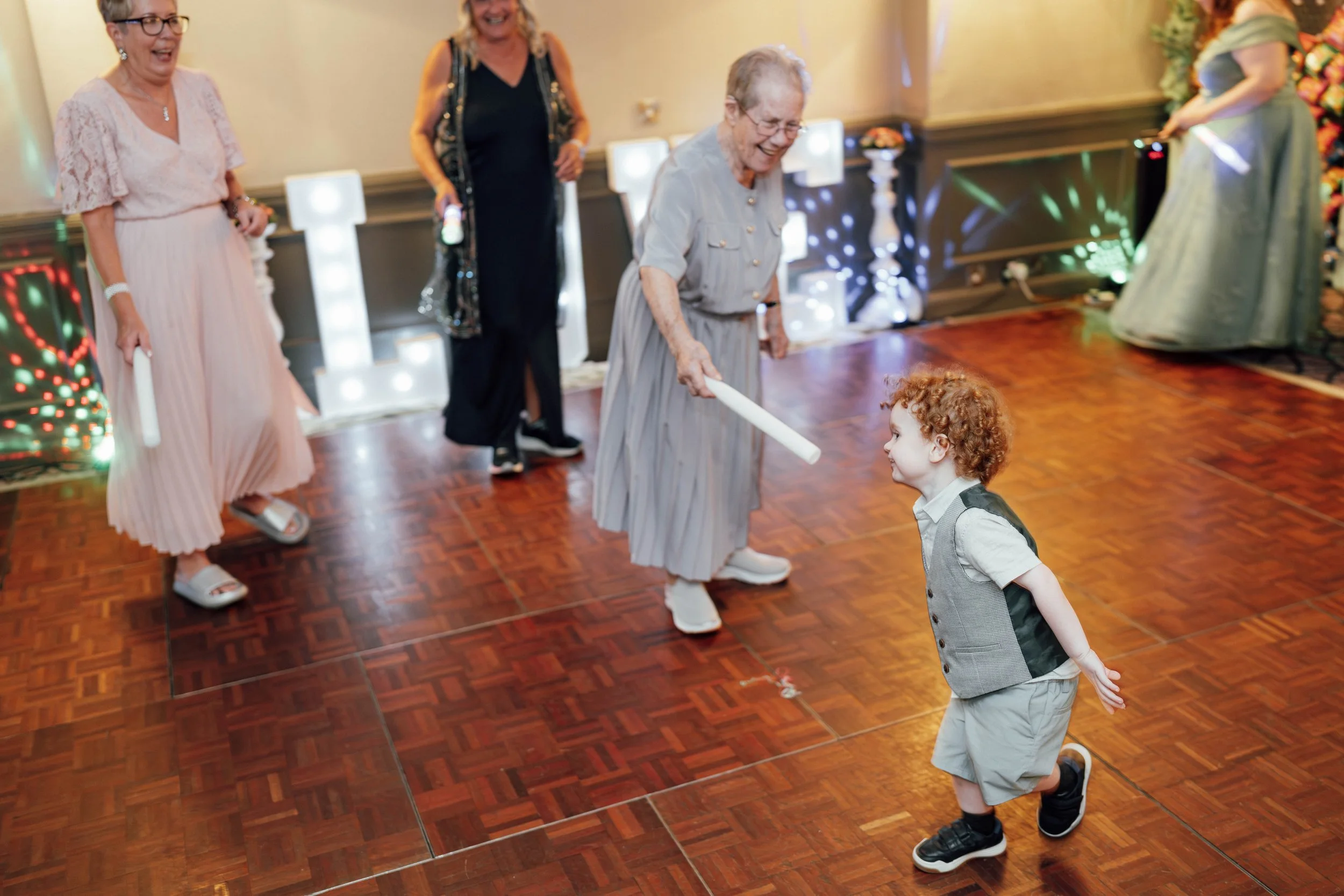 A young boy with curly red hair dancing with an elderly woman with short gray hair at a party. The woman is holding a glow stick and smiling, while the boy is in mid-dance with a bright smile. There are three other women in the background, two holdin