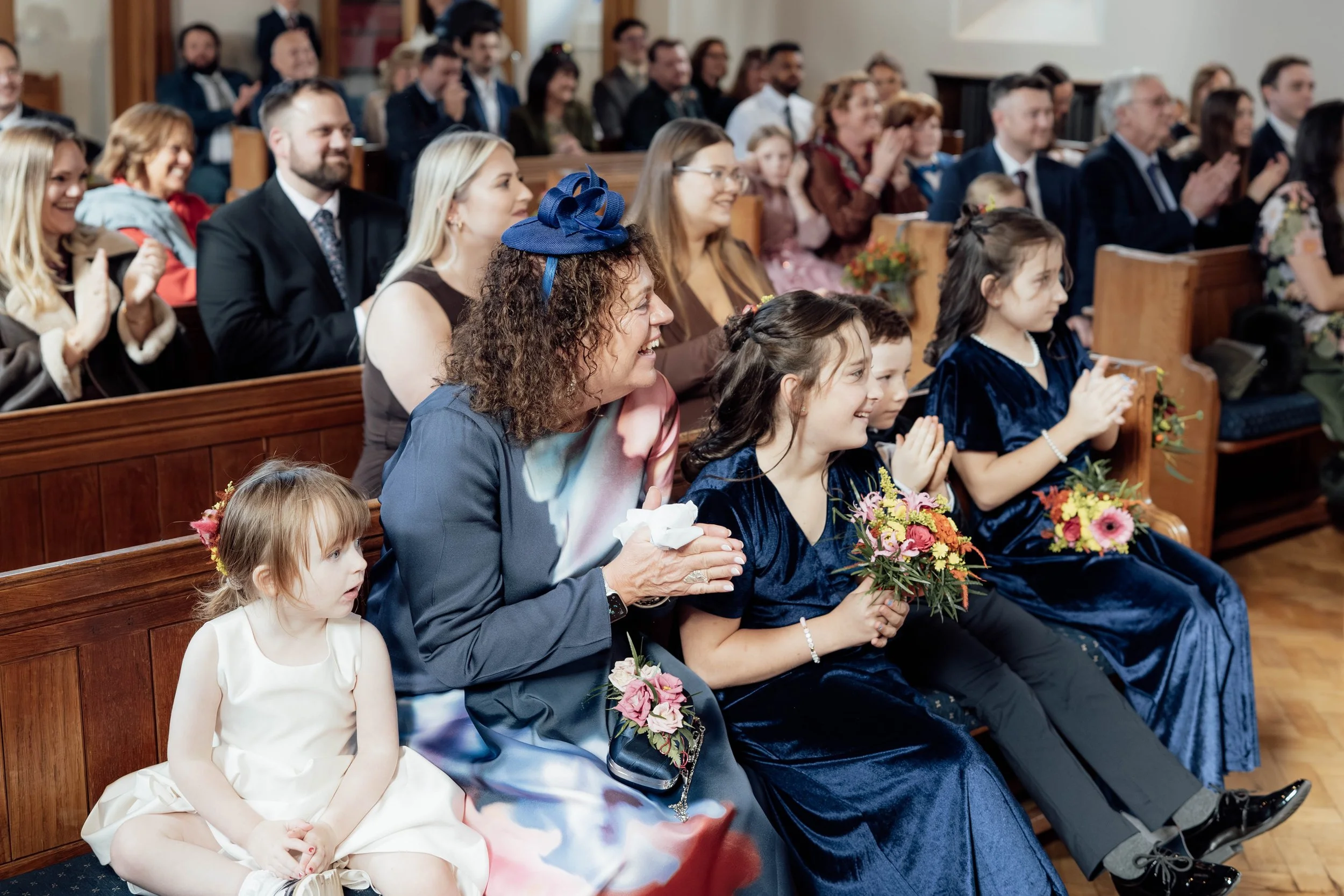 A group of children and adults attending a formal event inside a church, with some children holding bouquets of flowers, and many adults clapping and smiling.