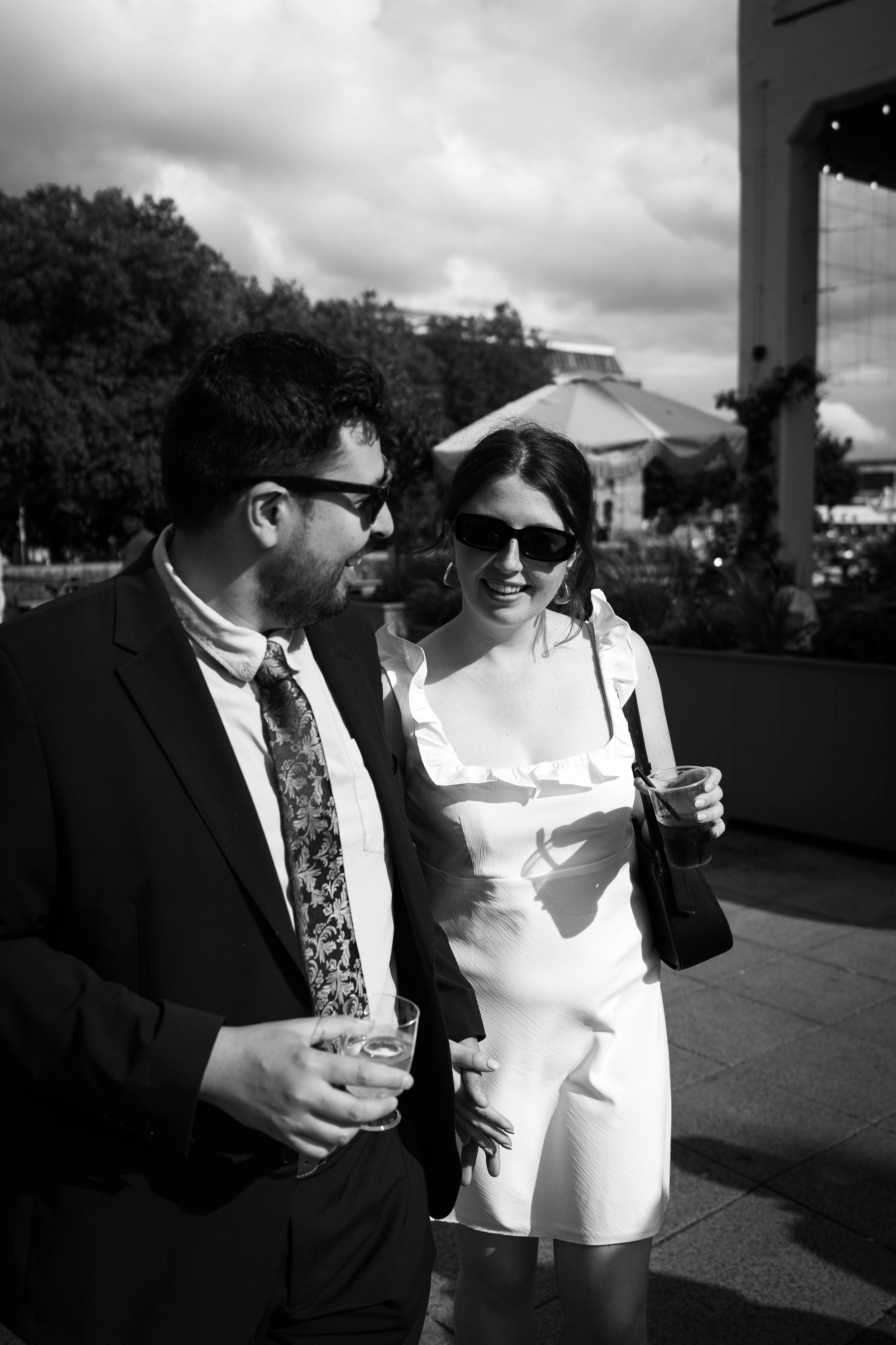 Two people, a man and a woman, wearing sunglasses and smiling, standing outdoors at a social gathering on a partly cloudy day.