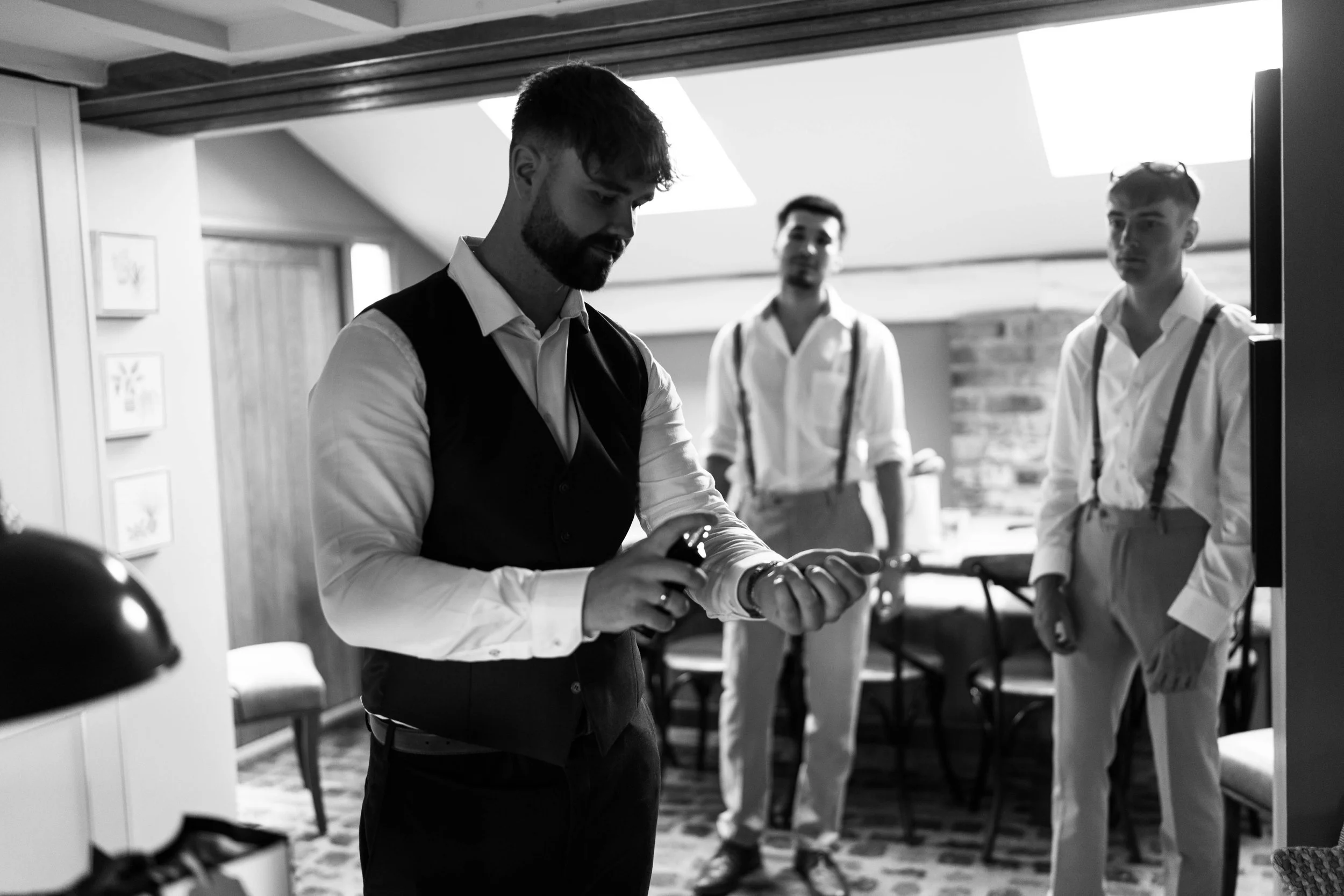 Three men in formal attire preparing for a special occasion in a cozy, well-lit room. One man in the foreground adjusts his cufflinks, while two others stand in the background observing.