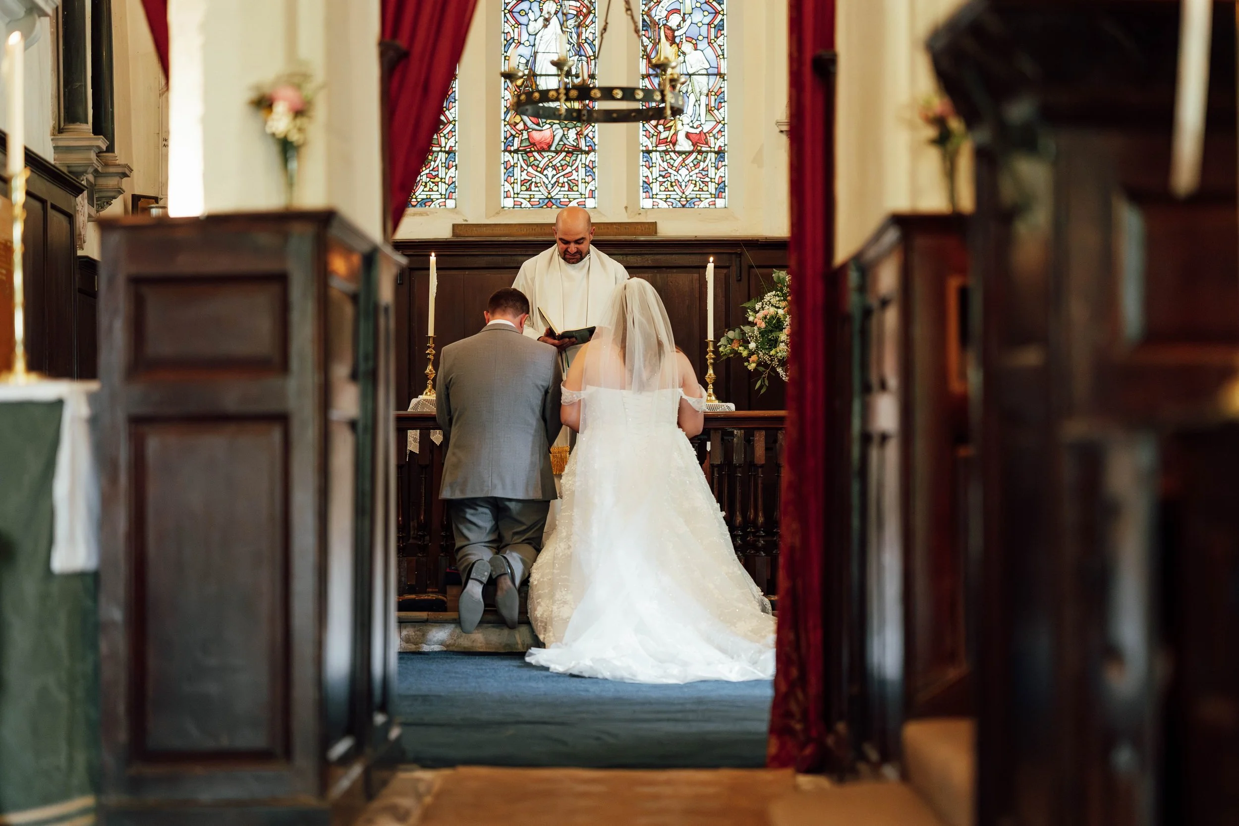 A bride and groom kneeling at the altar during a wedding ceremony in a church with stained glass windows, with a priest officiating.