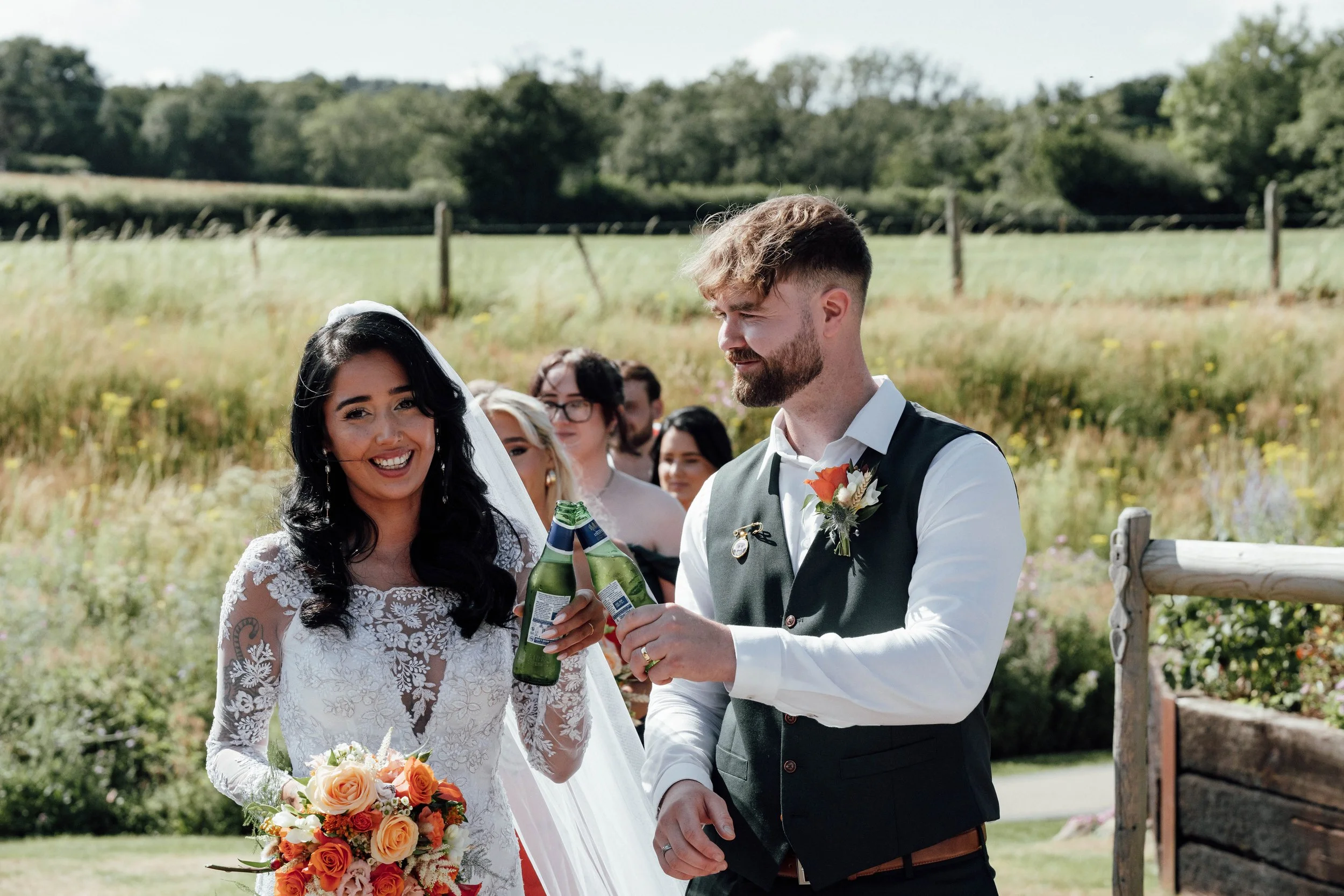 A bride and groom share a toast with drinks outdoors at their wedding, surrounded by friends on a sunny day in a grassy field.