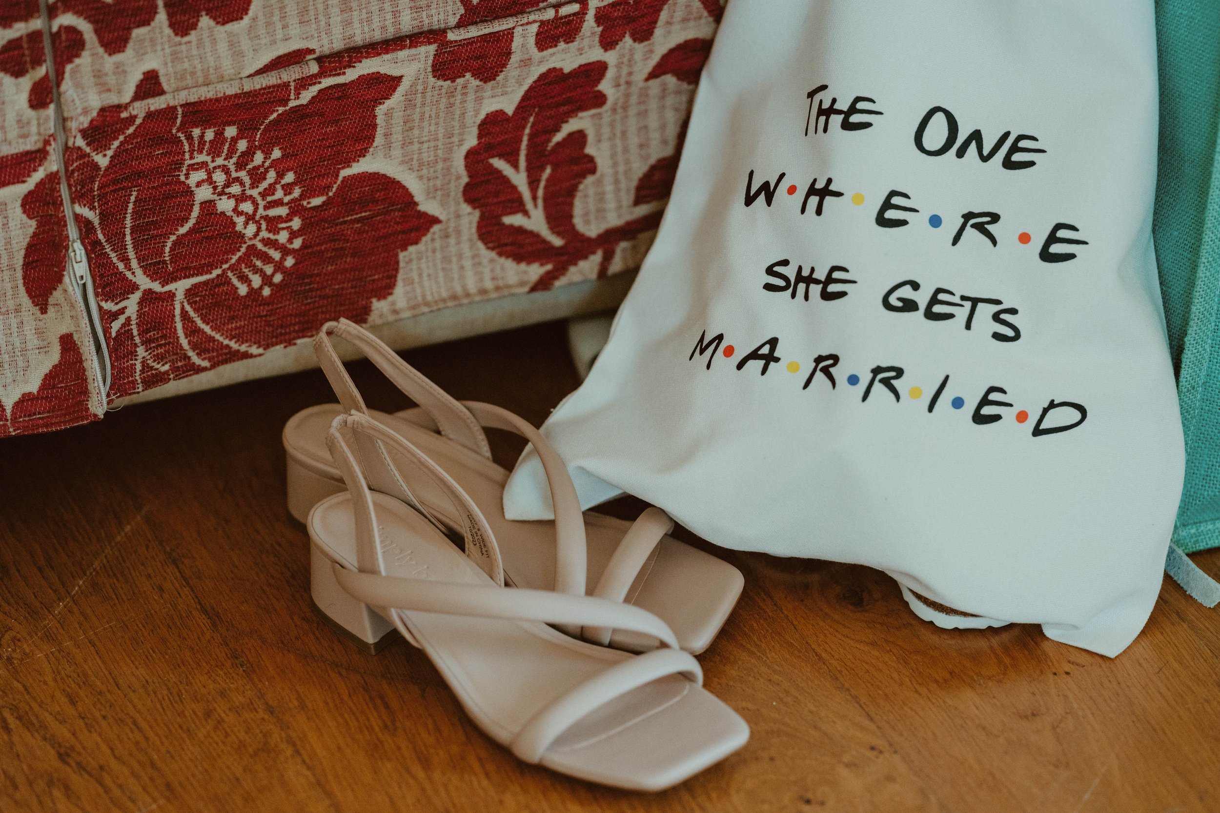 A pair of beige high-heeled strap sandals on a wooden floor, next to a white tote bag with the humorous quote, 'The one where she gets married,' referencing the TV show Friends, placed behind a floral-patterned fabric-covered furniture.