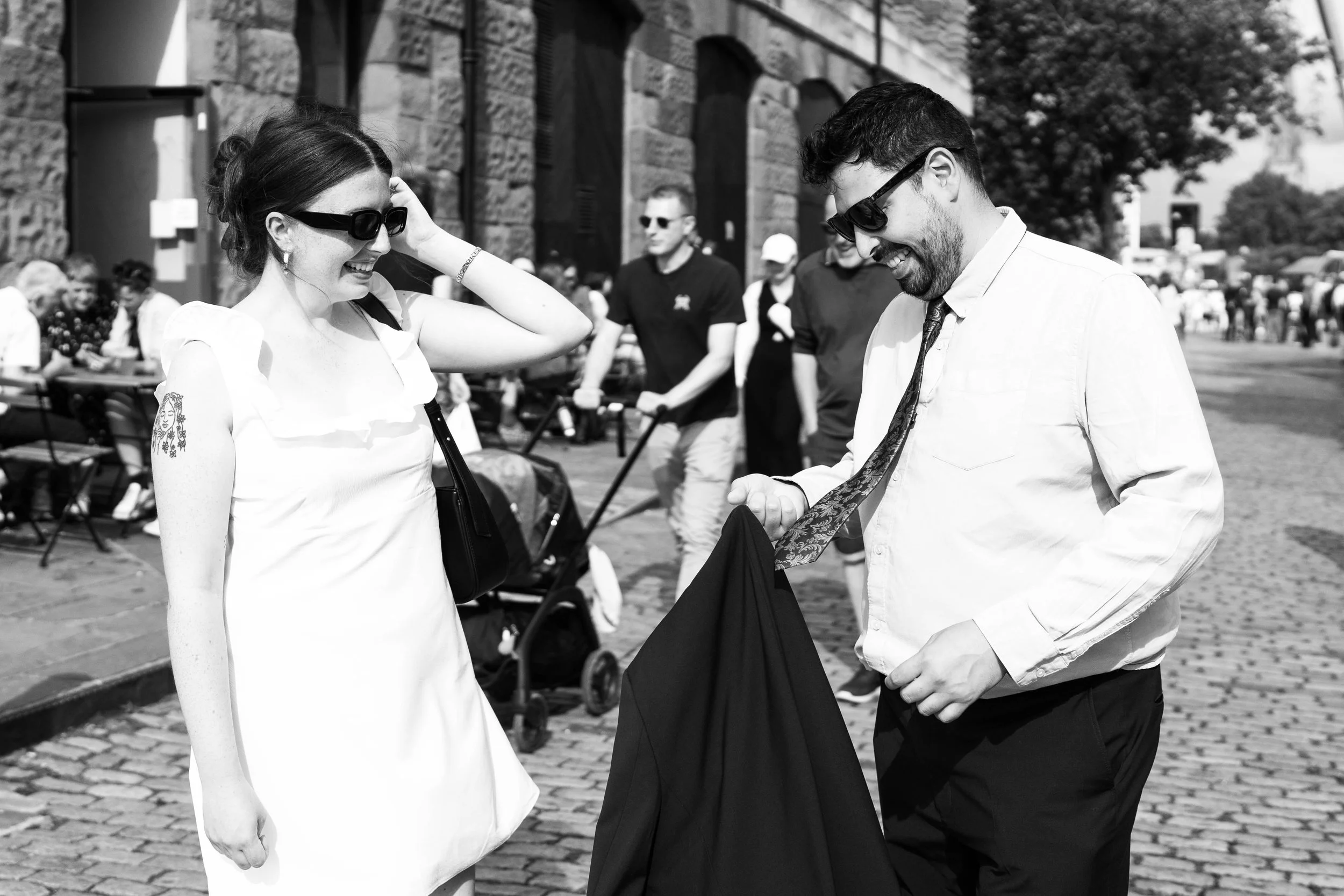 A woman and a man are smiling and talking outdoors on a sunny day, with other people and a stroller in the background.
