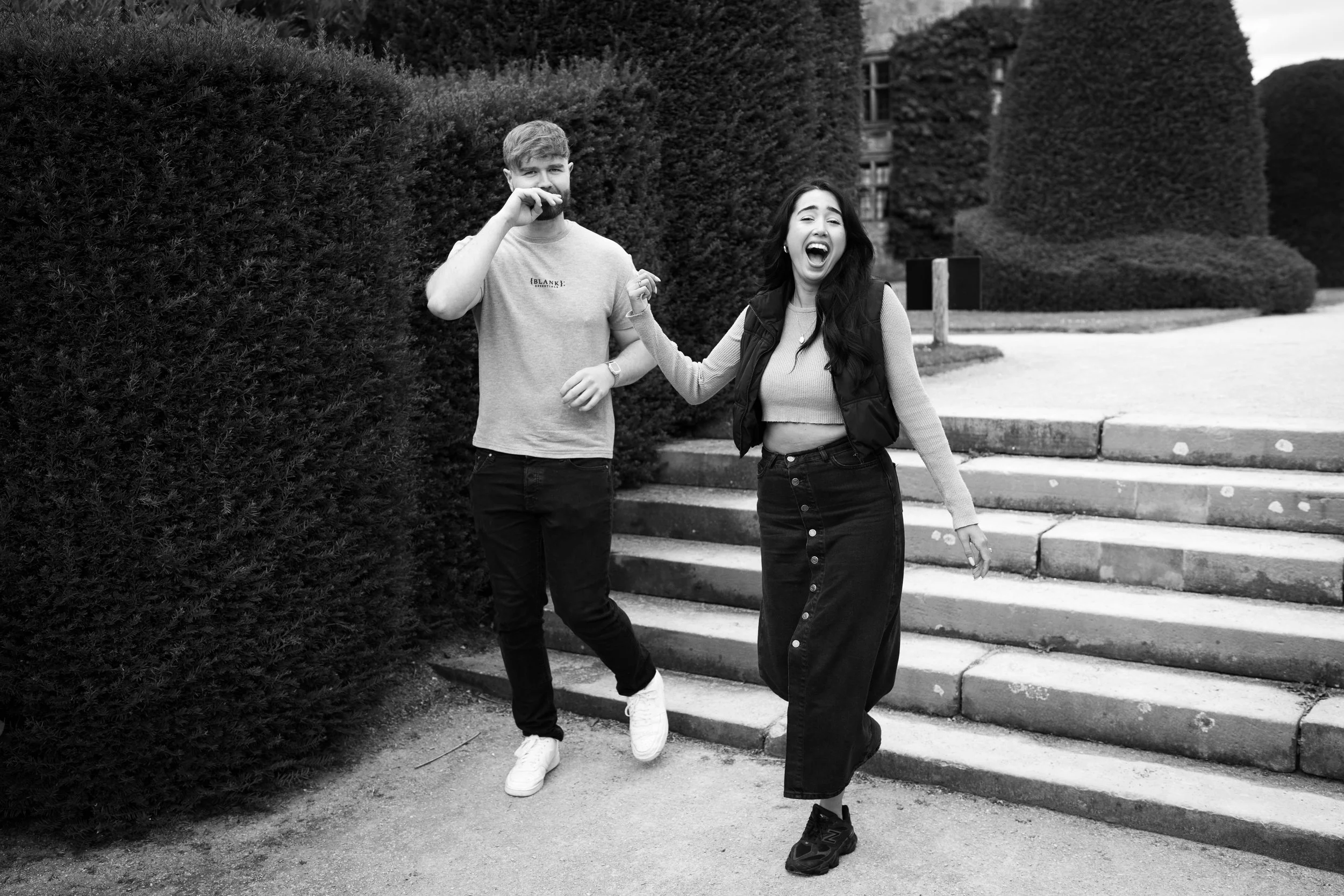 Two young people, a man and a woman, are outdoors on steps, laughing and smiling. The woman is holding the man's hand. The scene appears joyful and casual, with trees and a building in the background.