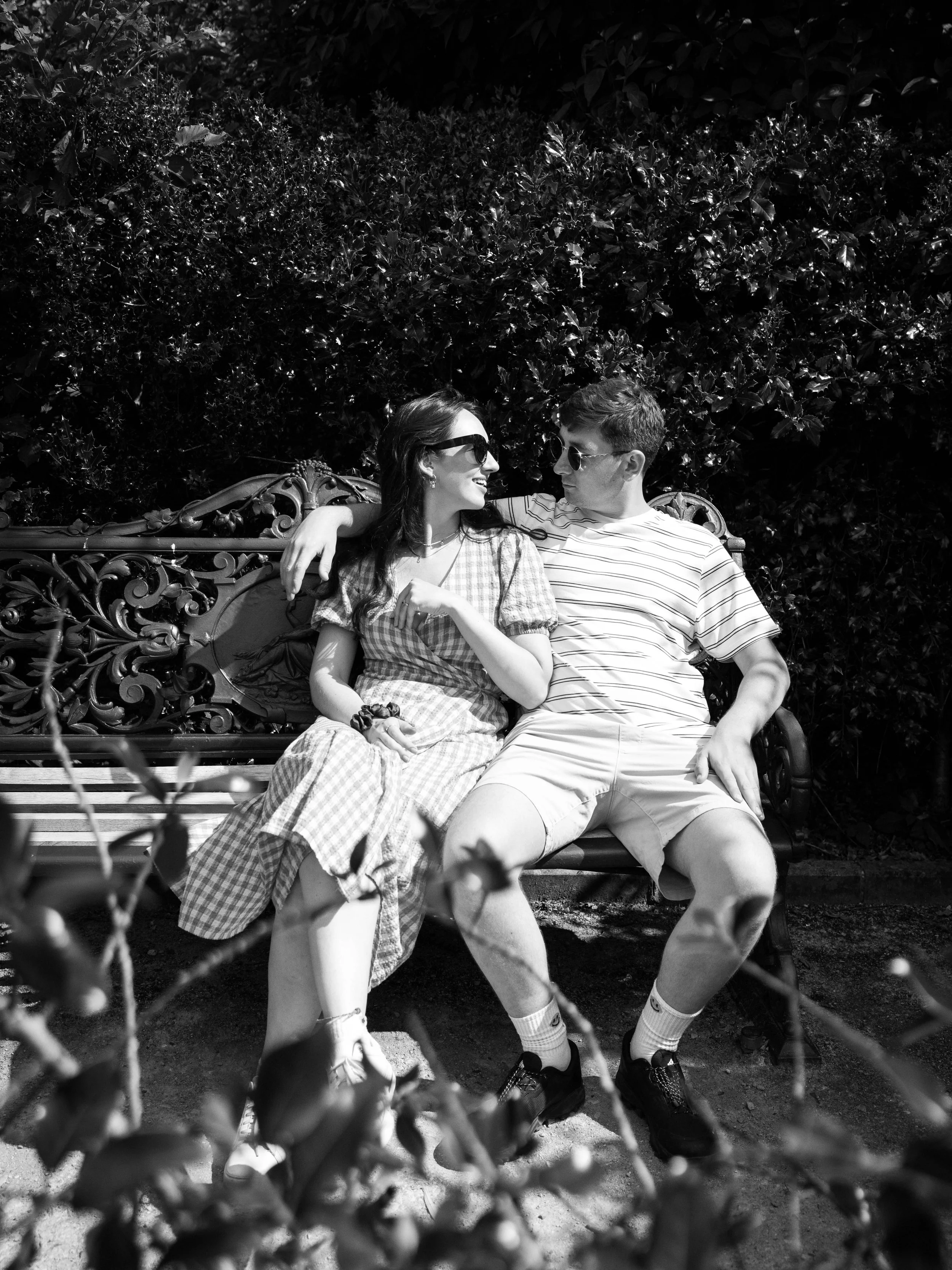 A black-and-white photo of a young couple sitting close and smiling on a decorative park bench, surrounded by foliage.