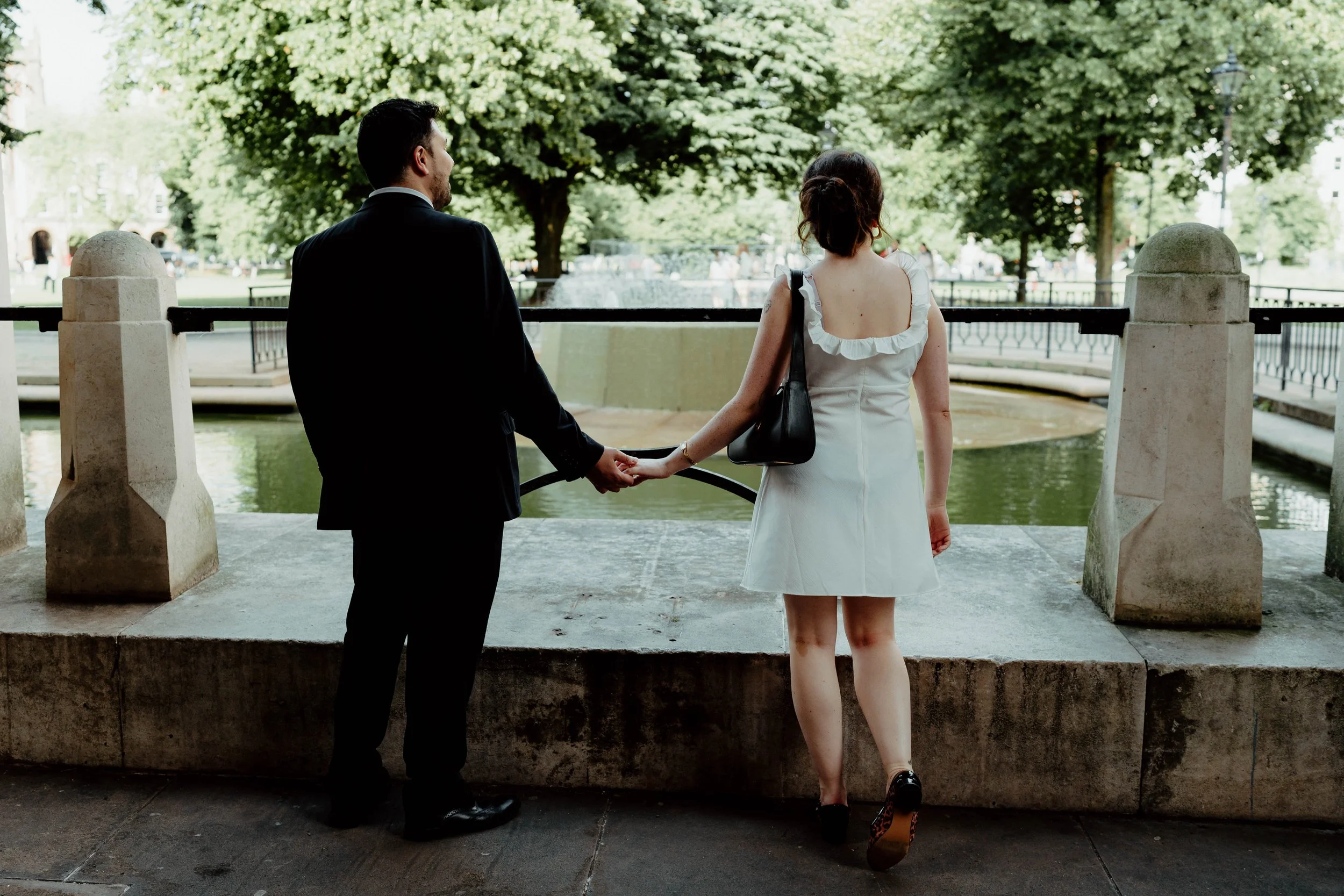A man and woman holding hands while standing by a fountain in a park with trees in the background.