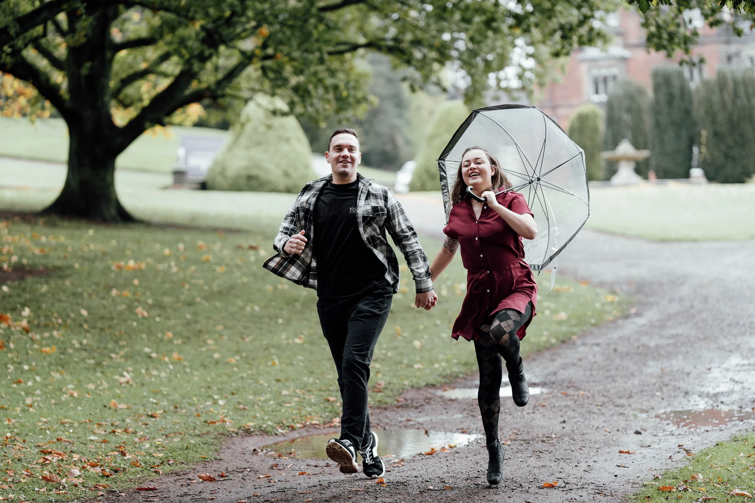 A smiling man and woman holding hands and running outdoors on a park trail with trees and grass, the woman holding a clear umbrella.