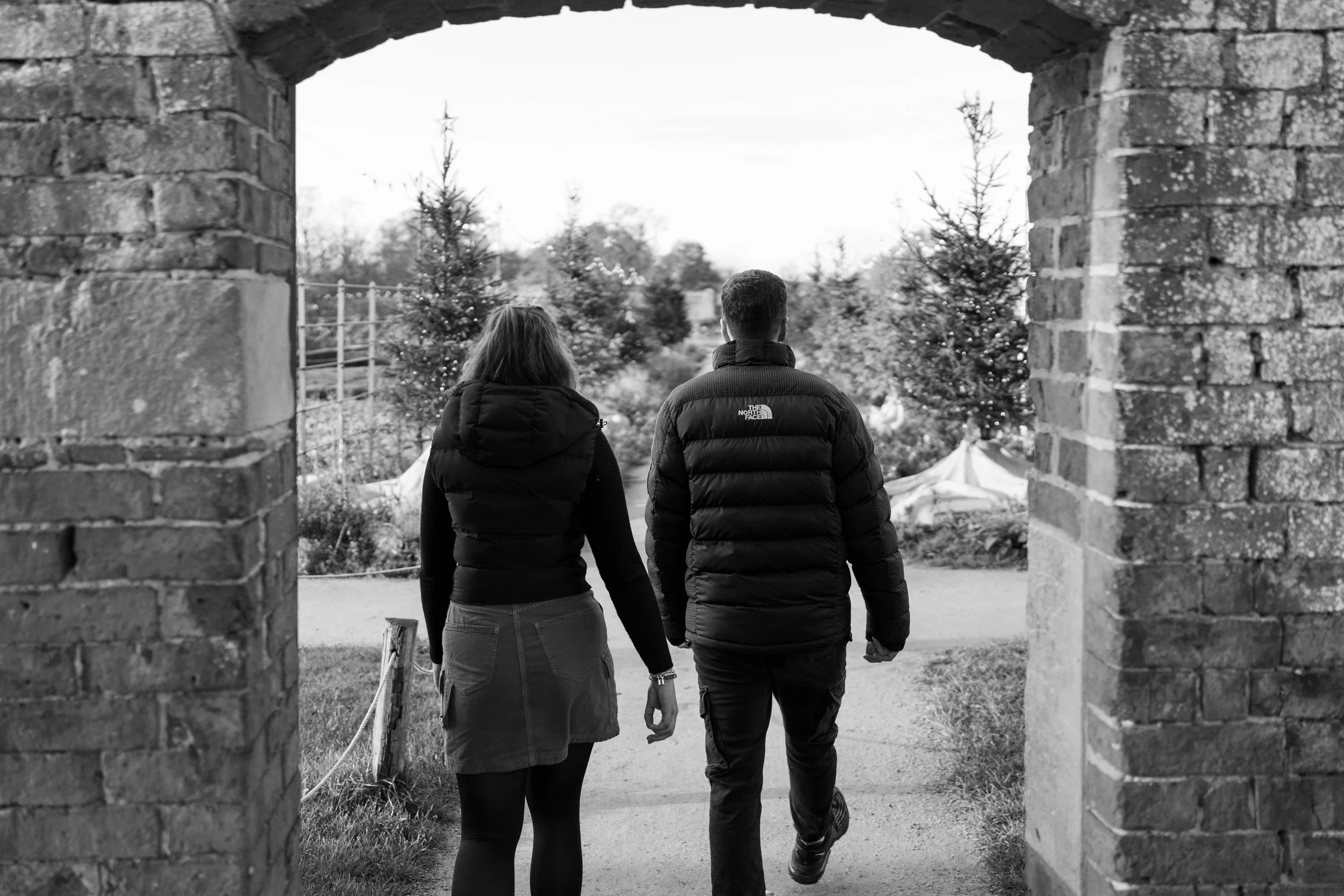 A black-and-white photo of a man and woman walking hand in hand through an arched brick gateway towards a Christmas tree with lights in an outdoor setting.