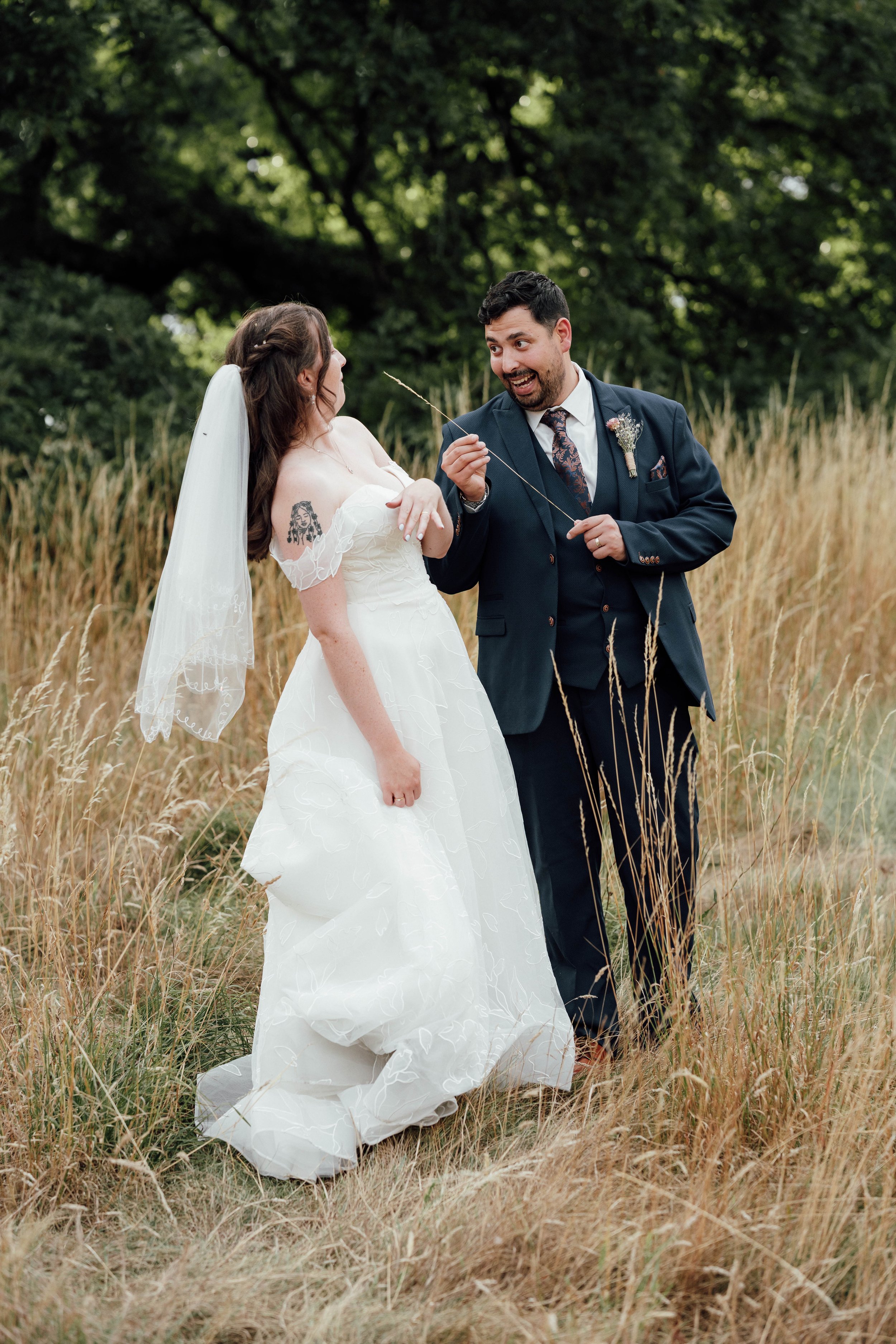 A bride and groom in wedding attire standing outdoors in a field of tall grass, with the groom holding a small plant and the bride smiling.