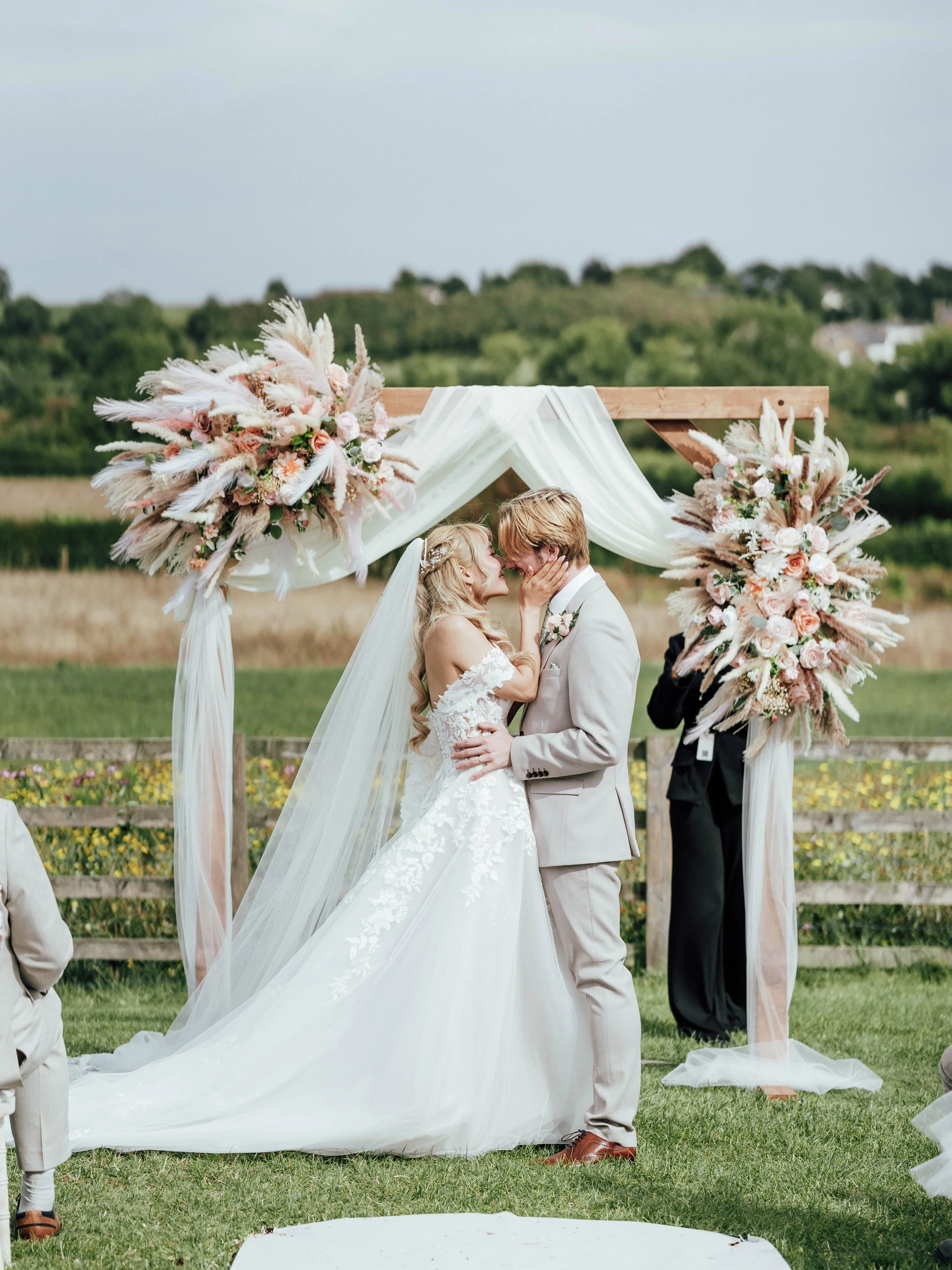 Bride and groom sharing a kiss during an outdoor wedding ceremony, standing under a wooden arch decorated with pink and white floral arrangements and draped fabric, with a green landscape background.
