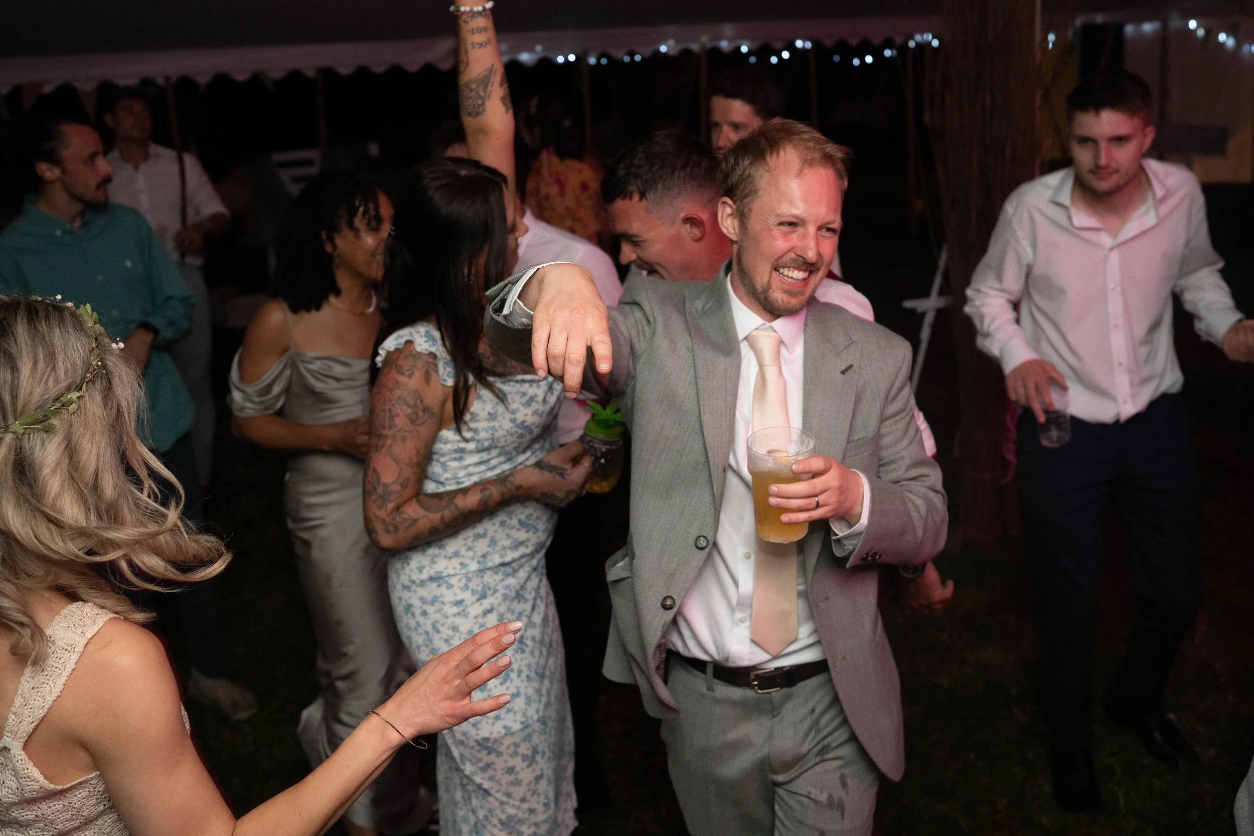 People dancing and socializing at a party or celebration, with a man in a gray suit holding a drink and smiling.