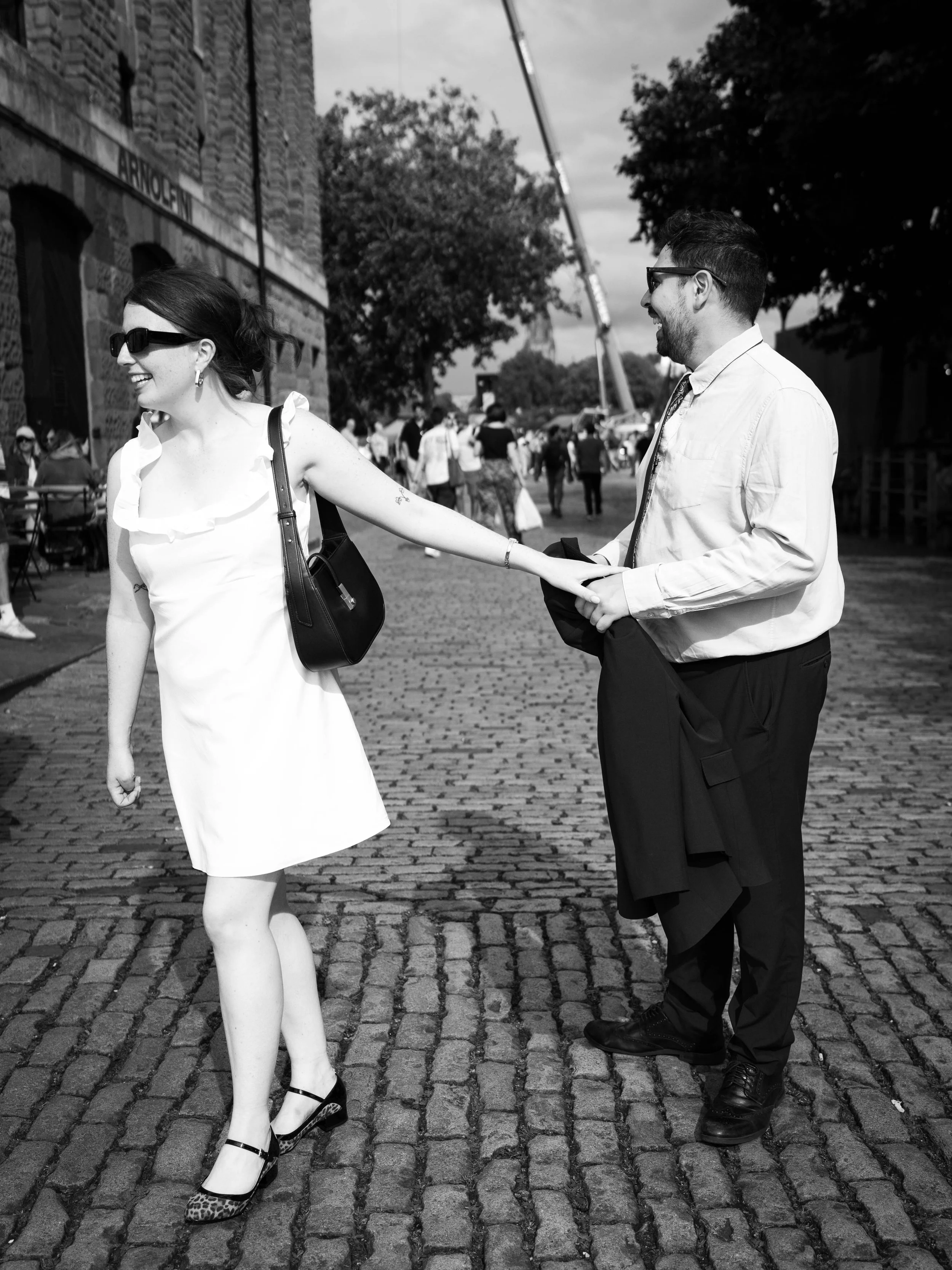 A woman in a white dress and sunglasses smiling and holding hands with a man in a white shirt, black trousers, and sunglasses on a cobblestone street, with people and trees in the background.