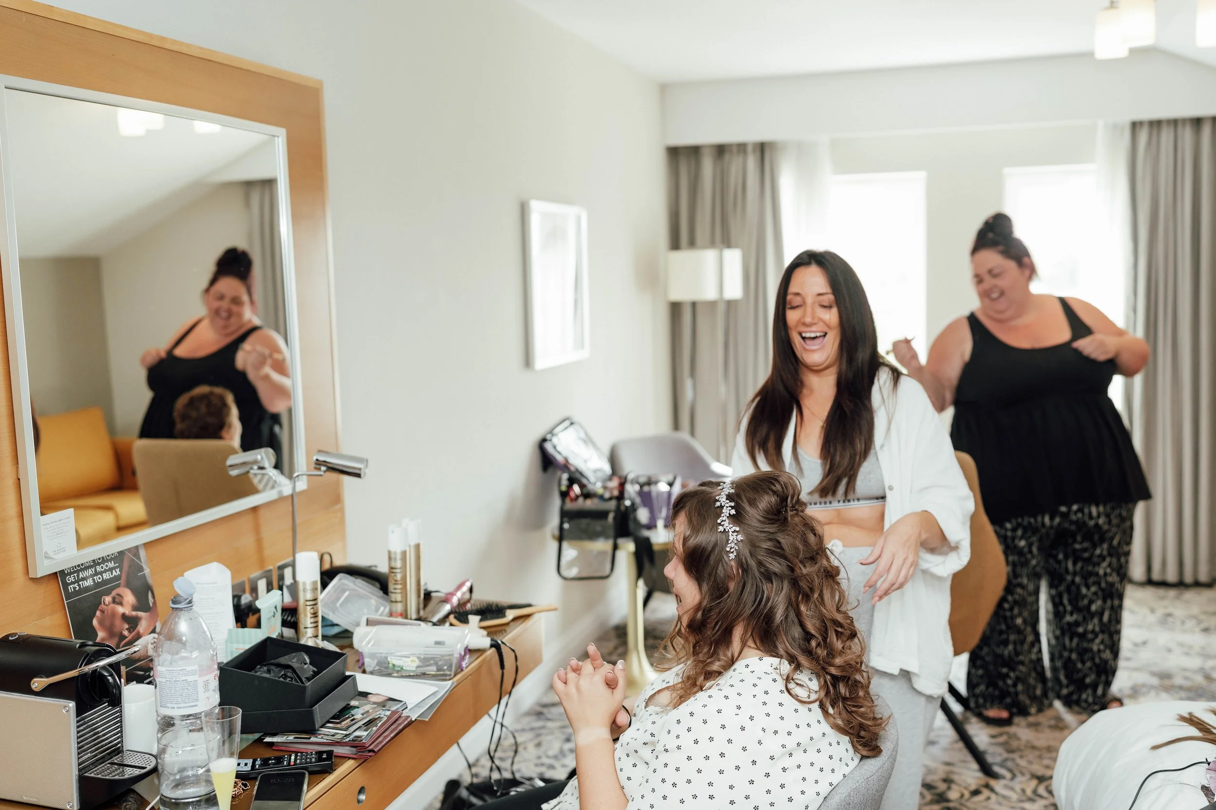 Two women in front of a mirror with one applying makeup and the other with a tiara, while another woman in black with a bun hairstyle dances in the background.