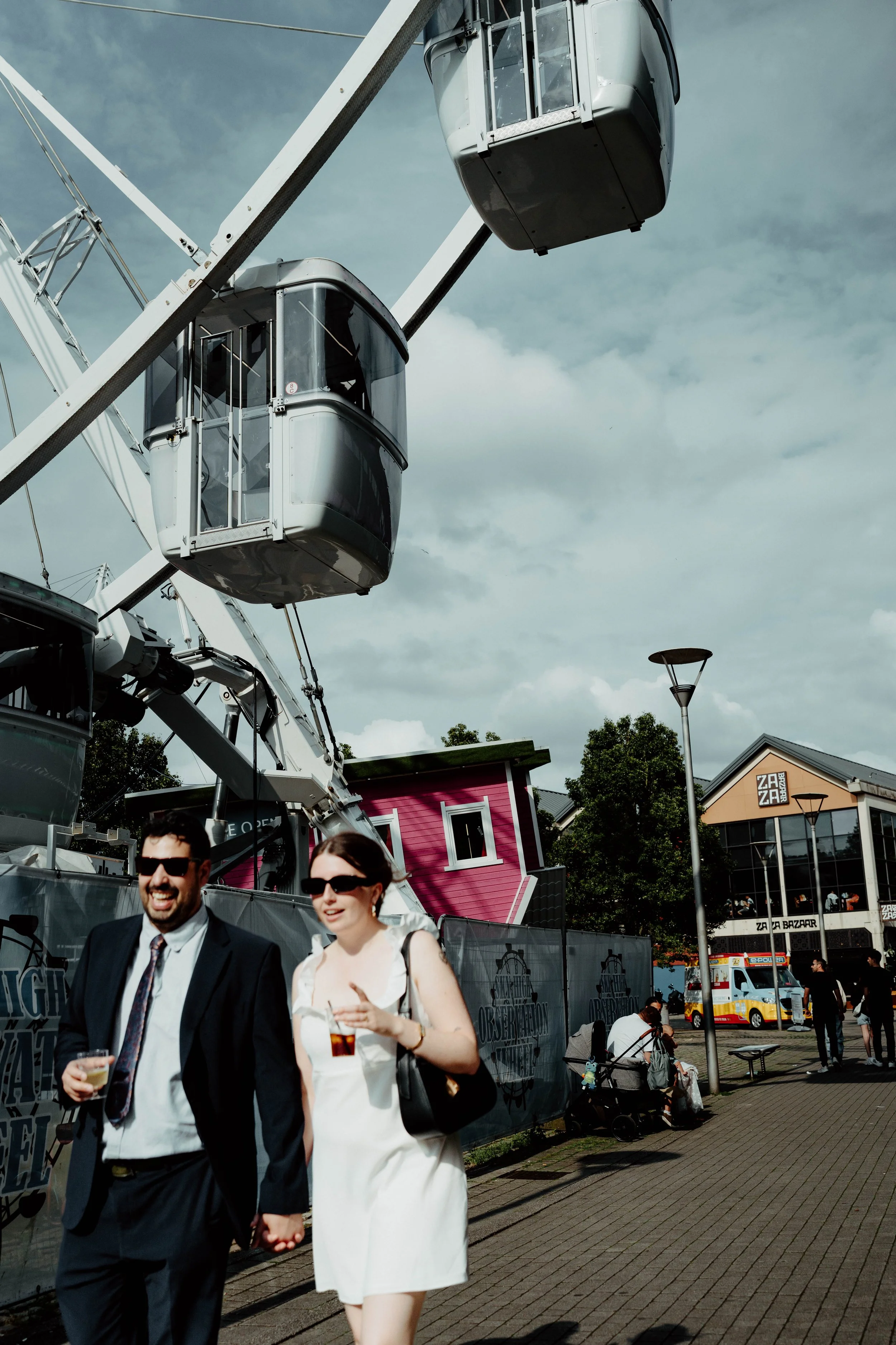 A ferris wheel with enclosed cabins at an outdoor fair, with a pink building, people sitting and walking, and an ice cream truck in the background.