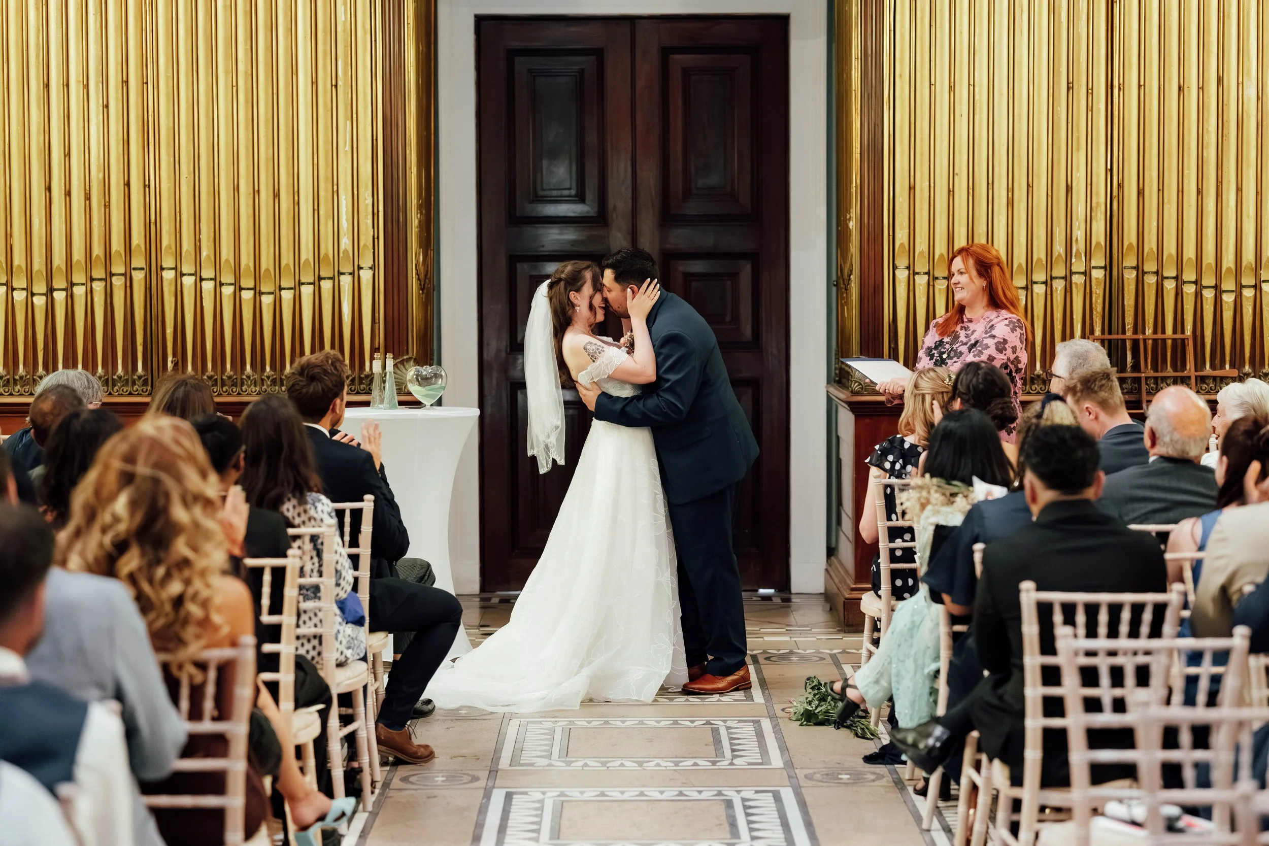 A couple kisses at their wedding ceremony in a decorated indoor venue, with guests seated on both sides and a wedding officiant standing to the right, holding a book.