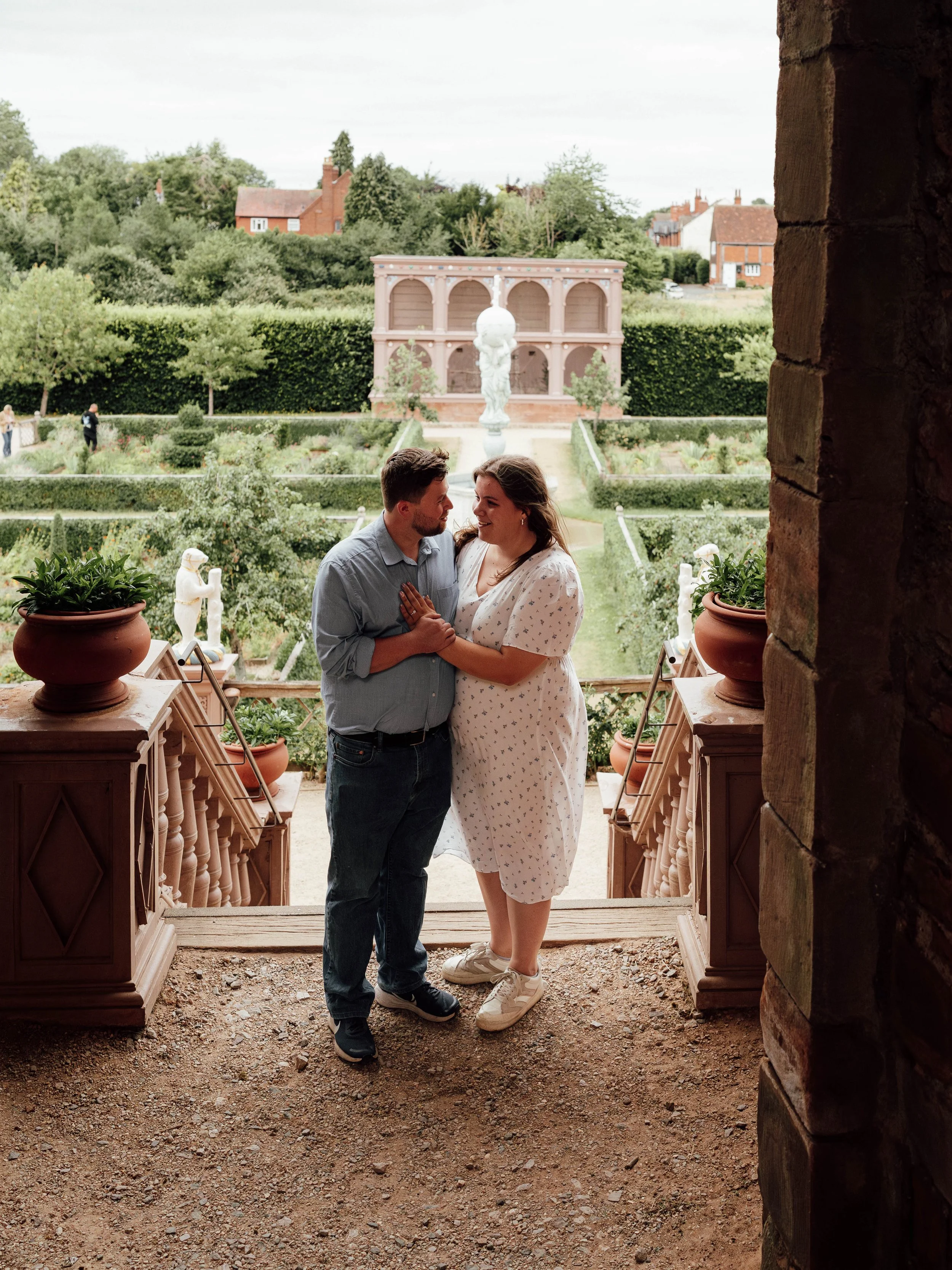 A couple stands on a small stone porch, facing each other and smiling, with a garden and an ornate fountain in the background.