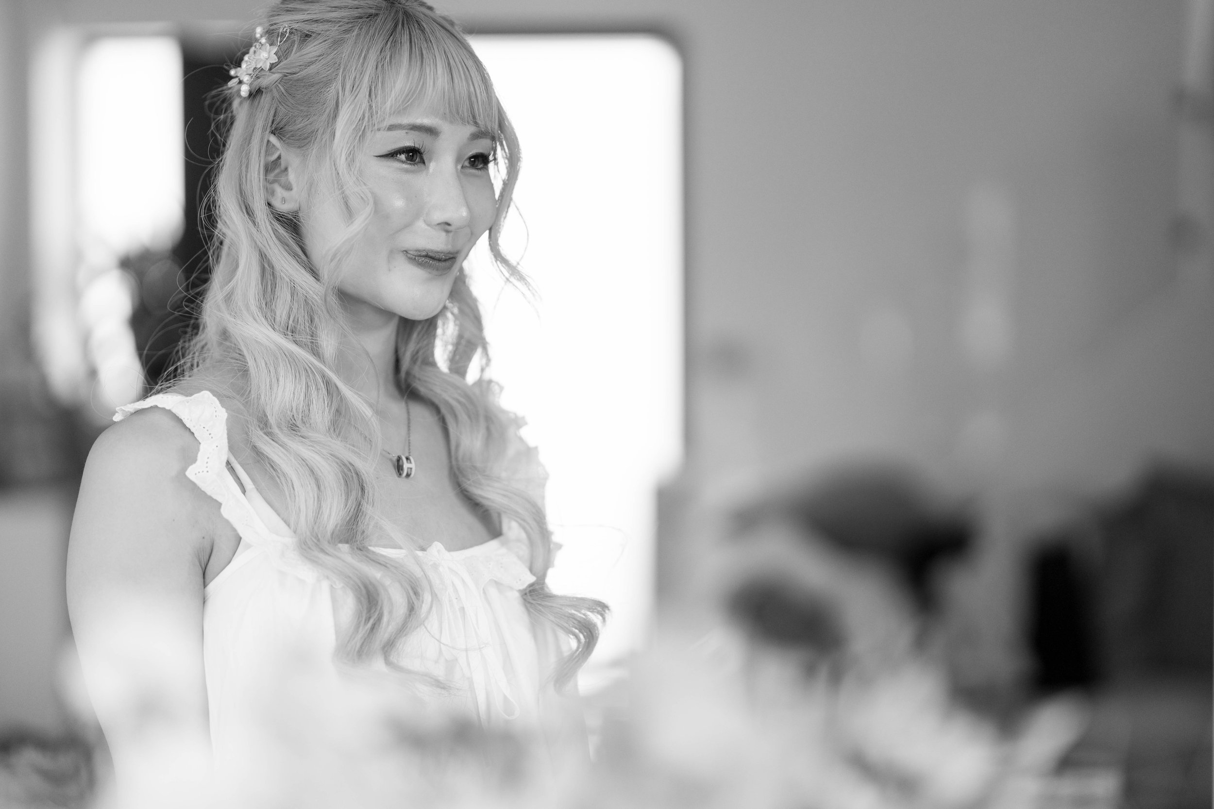 A young woman with long wavy hair adorned with a floral hair accessory, wearing a ruffled sleeveless top, smiling softly in a well-lit room.