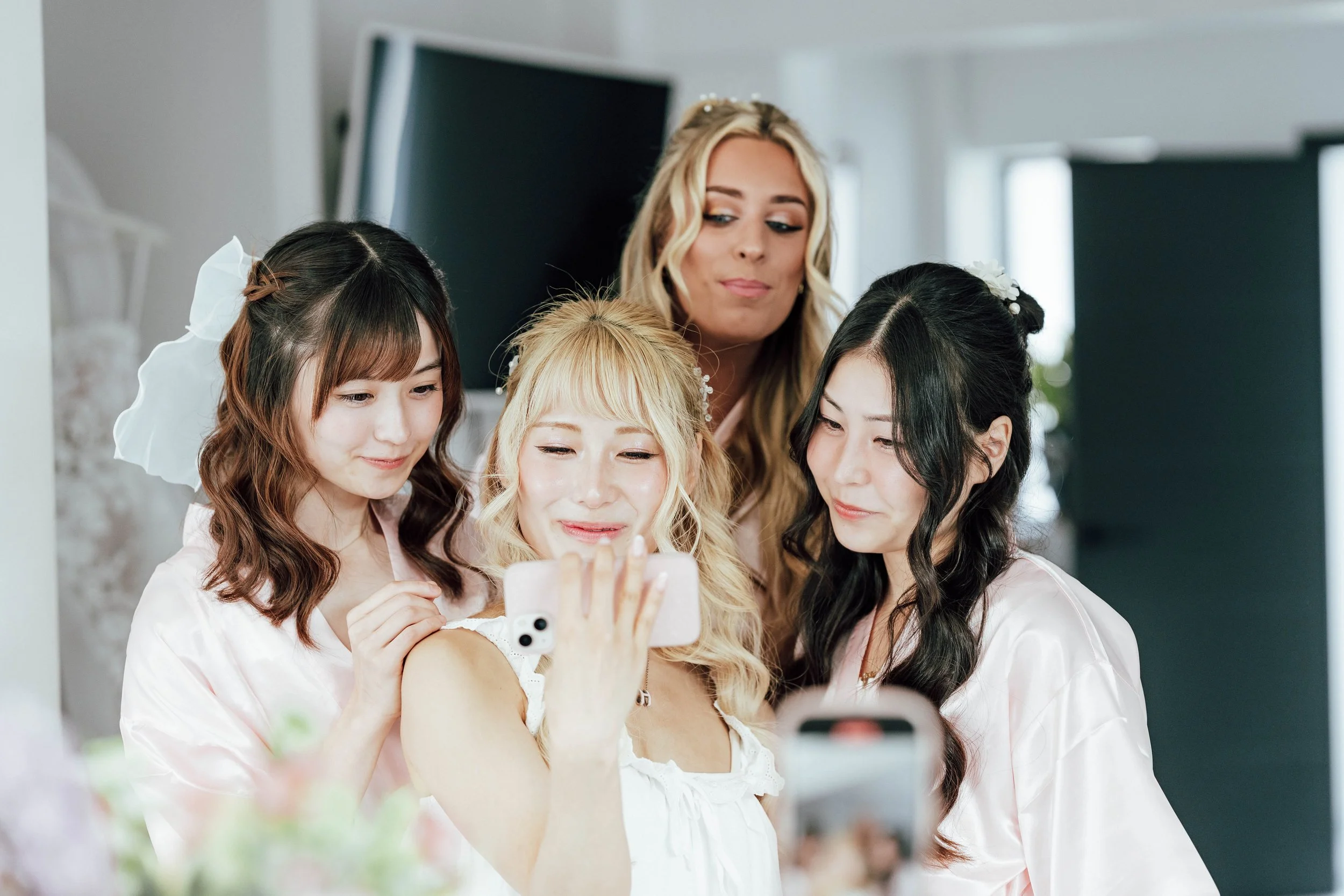 Four women, dressed in light pink and white robes, take a selfie together indoors. They are smiling and appear to be enjoying a special occasion, possibly preparing for a wedding.