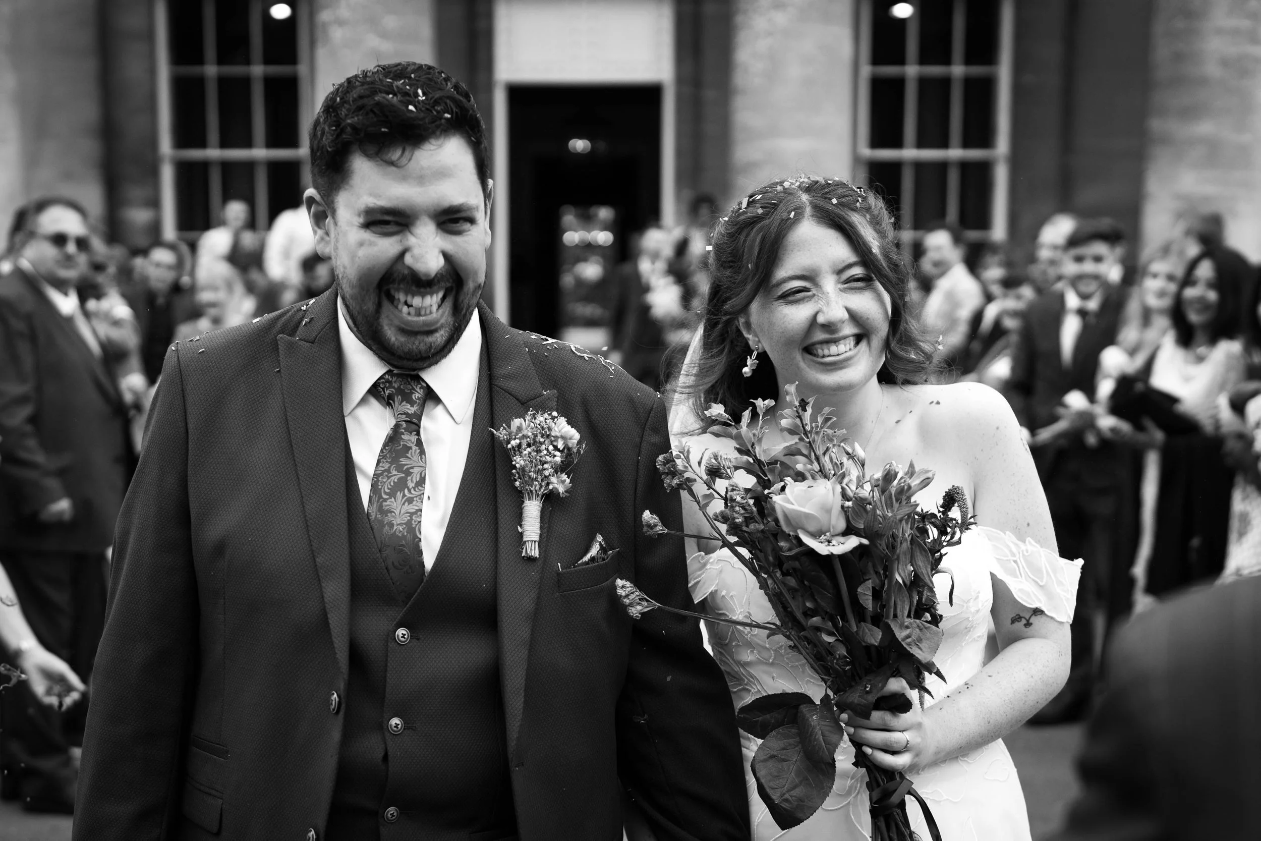 A black-and-white photo of a smiling bride and groom celebrating their wedding outdoors, surrounded by guests.
