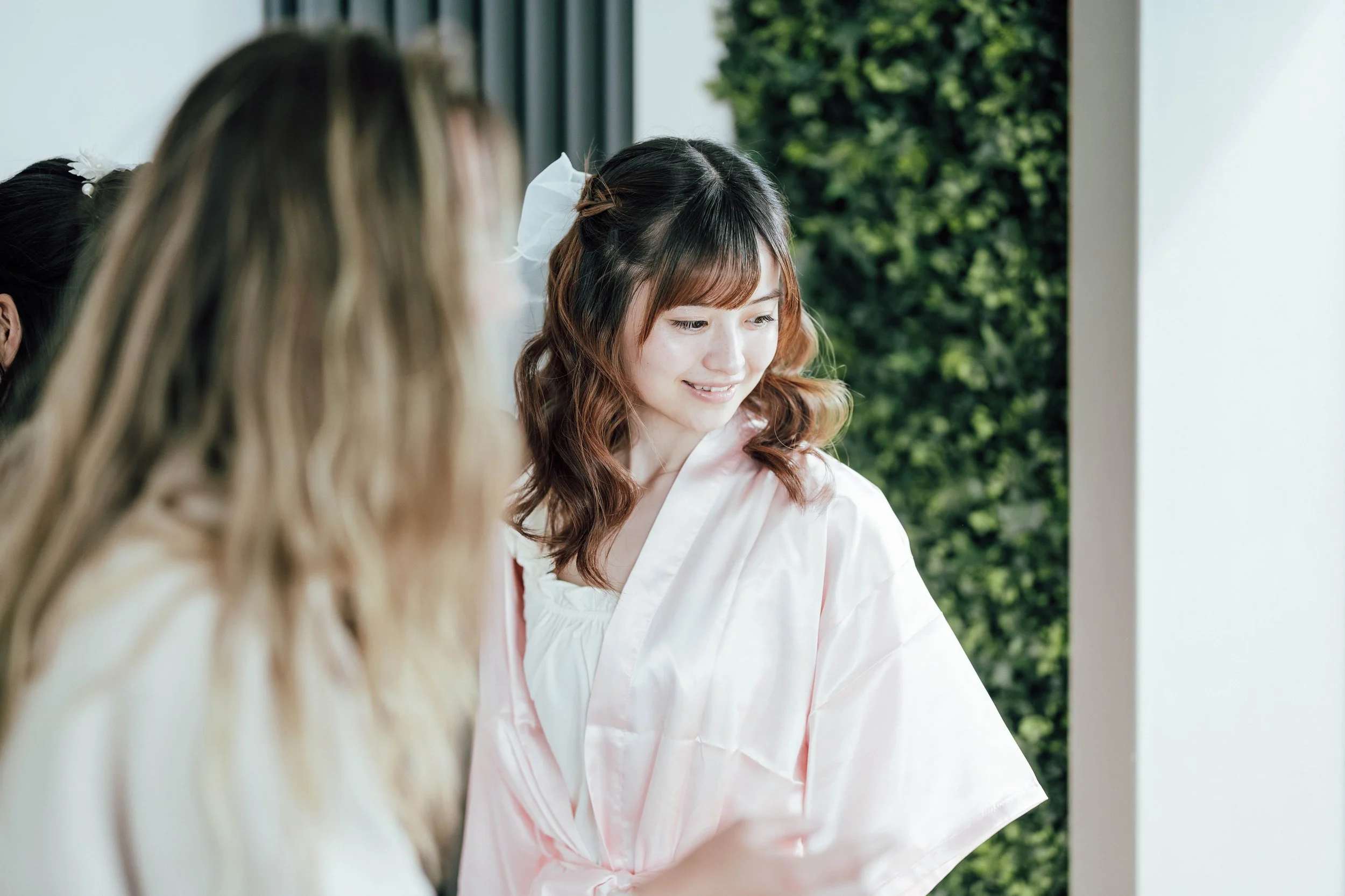 A woman in a light pink satin robe looking at her reflection in a mirror, with other women around her, in a bright room with green foliage outside.