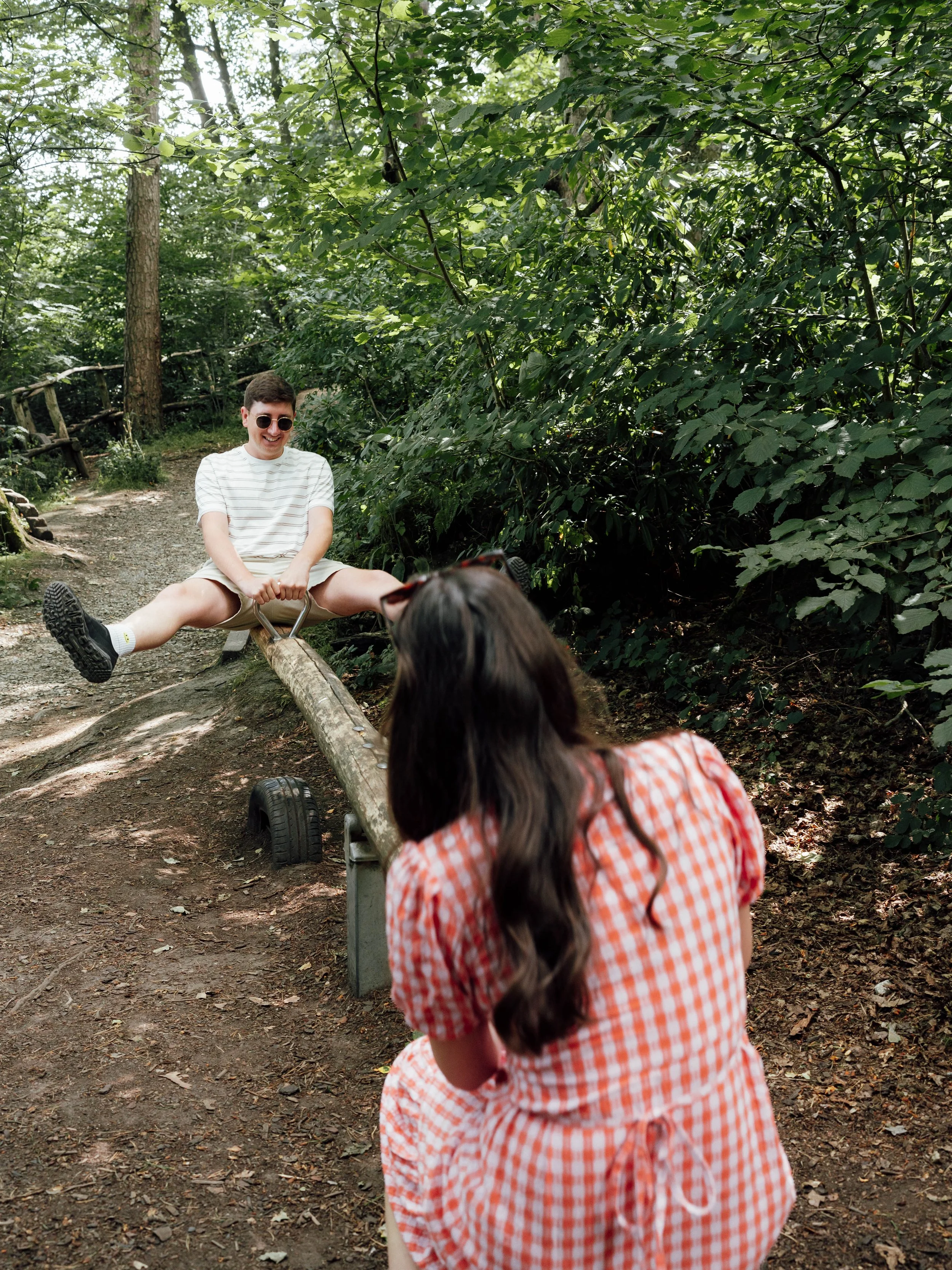 A young man and woman are playing on a seesaw in a wooded outdoor area. The young man, wearing sunglasses and a striped shirt, is sitting on one end with legs extended outward, while the woman, with long dark hair and a checkered dress, is sitting on