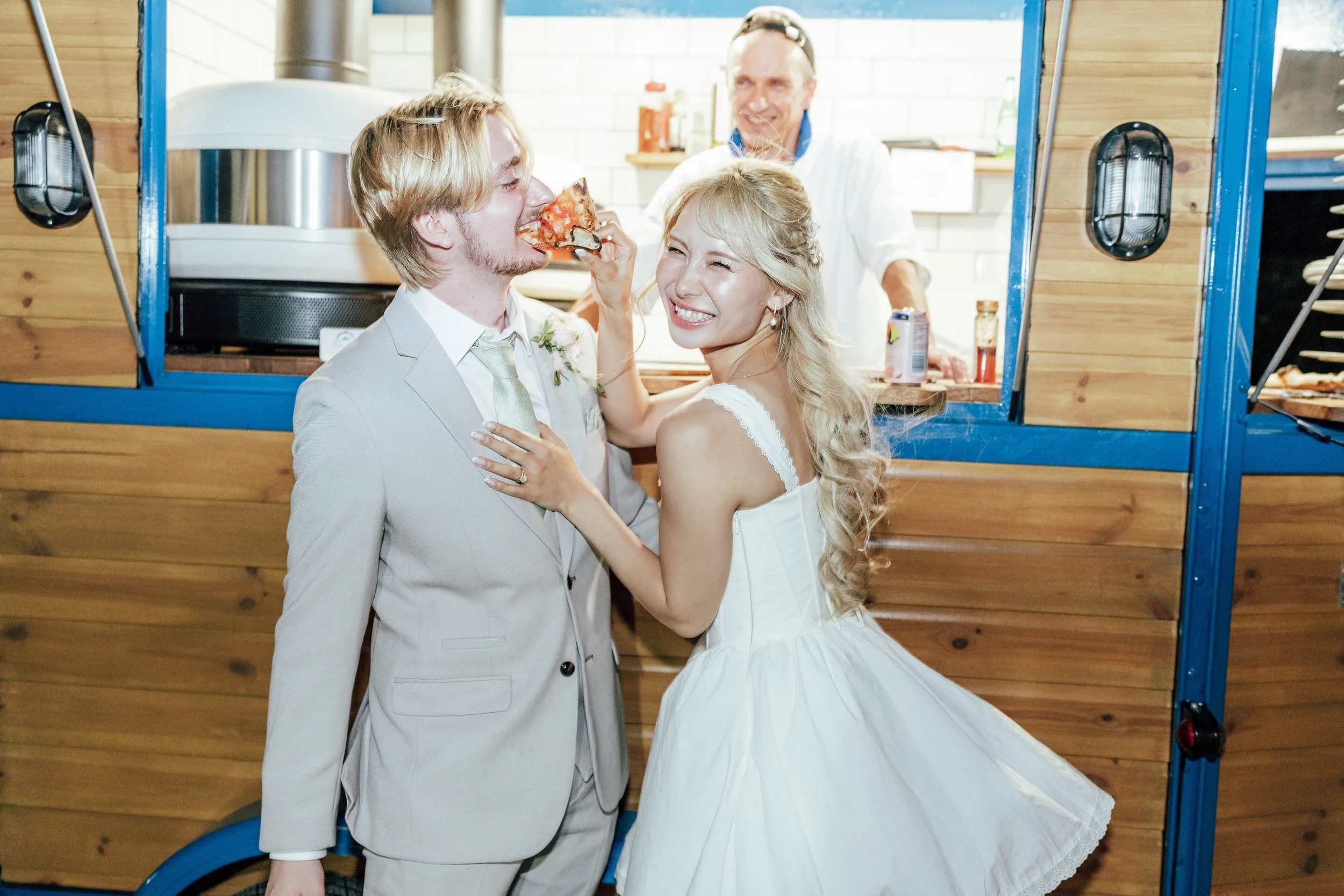 Bride and groom in wedding attire sharing a slice of pizza at a food truck, with a smiling man inside the truck.