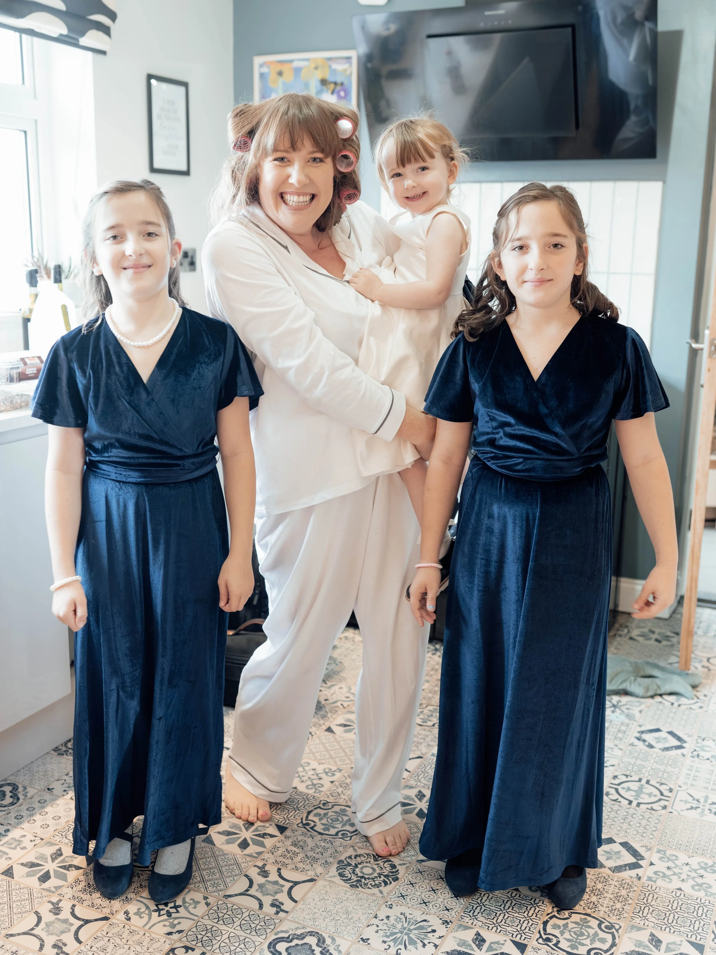 A woman with hair rollers in pajamas is holding a young girl in a white dress, surrounded by three girls in navy blue dresses, in a kitchen with patterned floor tiles and a wall-mounted TV.