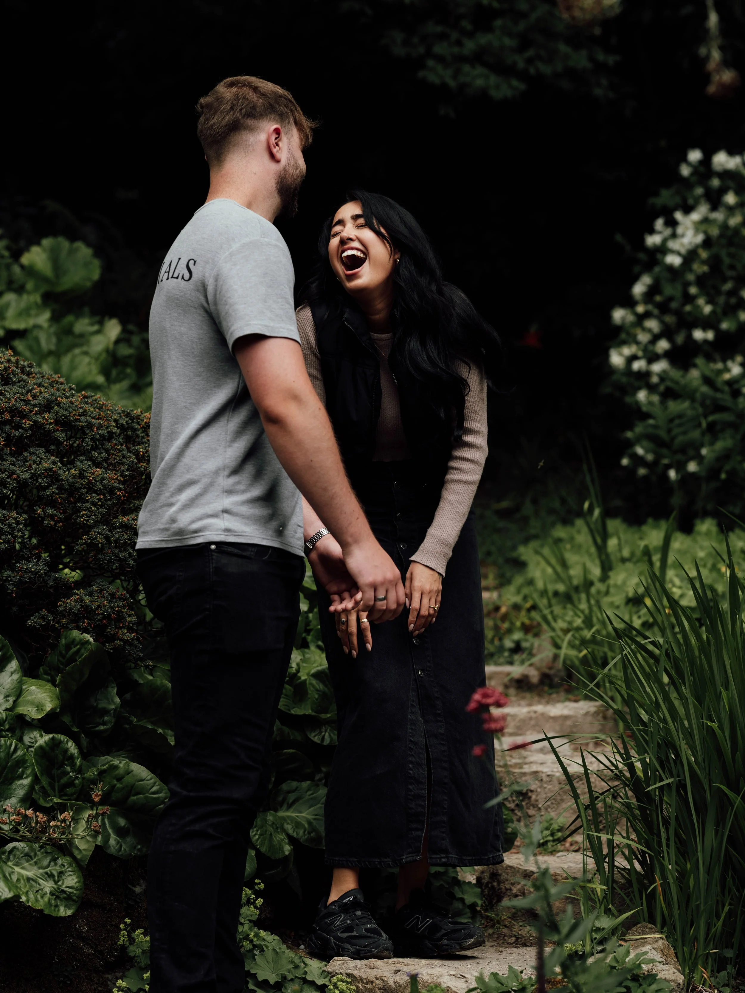 A young couple standing outdoors, holding hands, and laughing together in a lush garden.