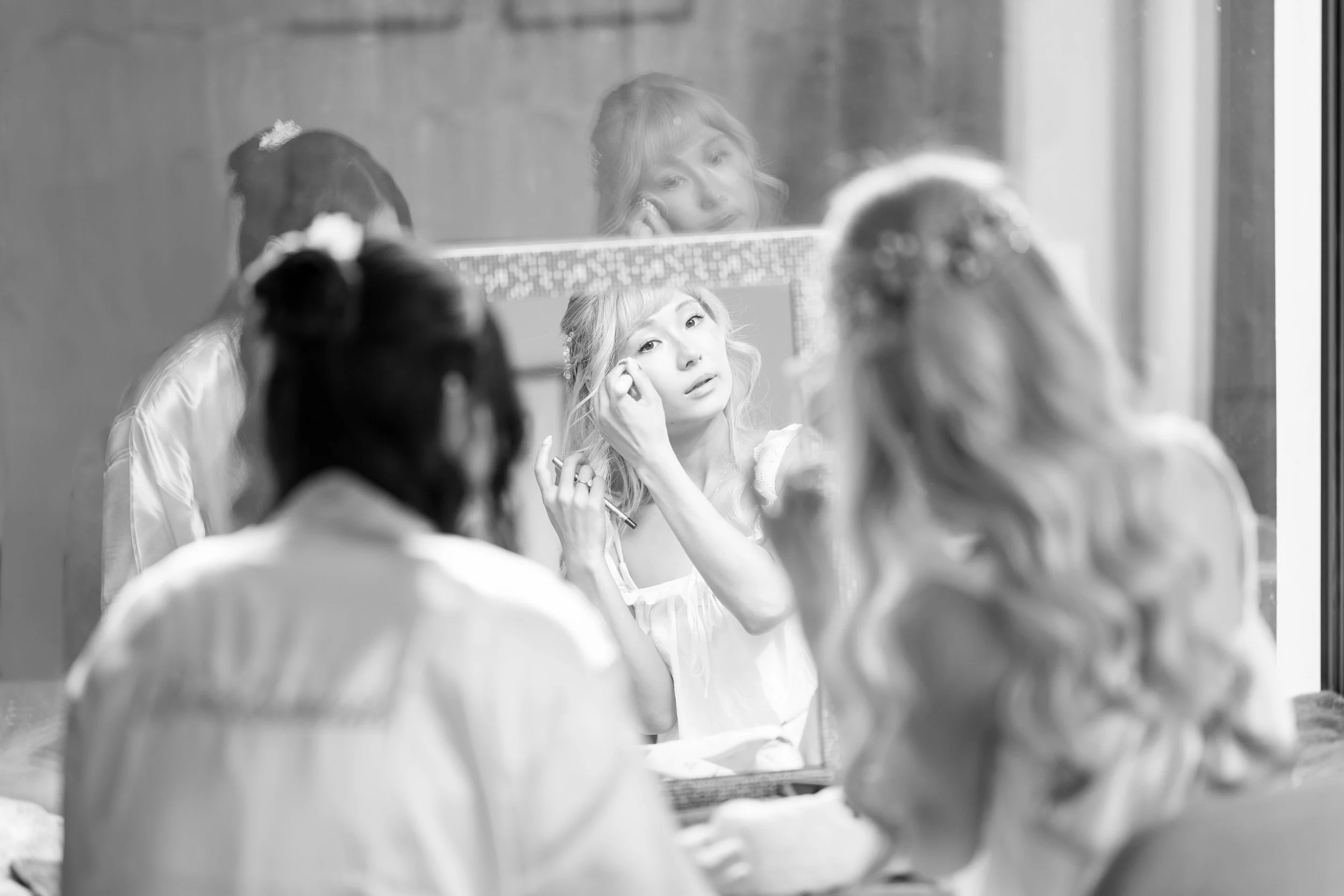 A woman is applying makeup in front of a mirror. She is surrounded by three other women, all with curly hair, two of whom have flowers in their hair. The scene is in black and white.