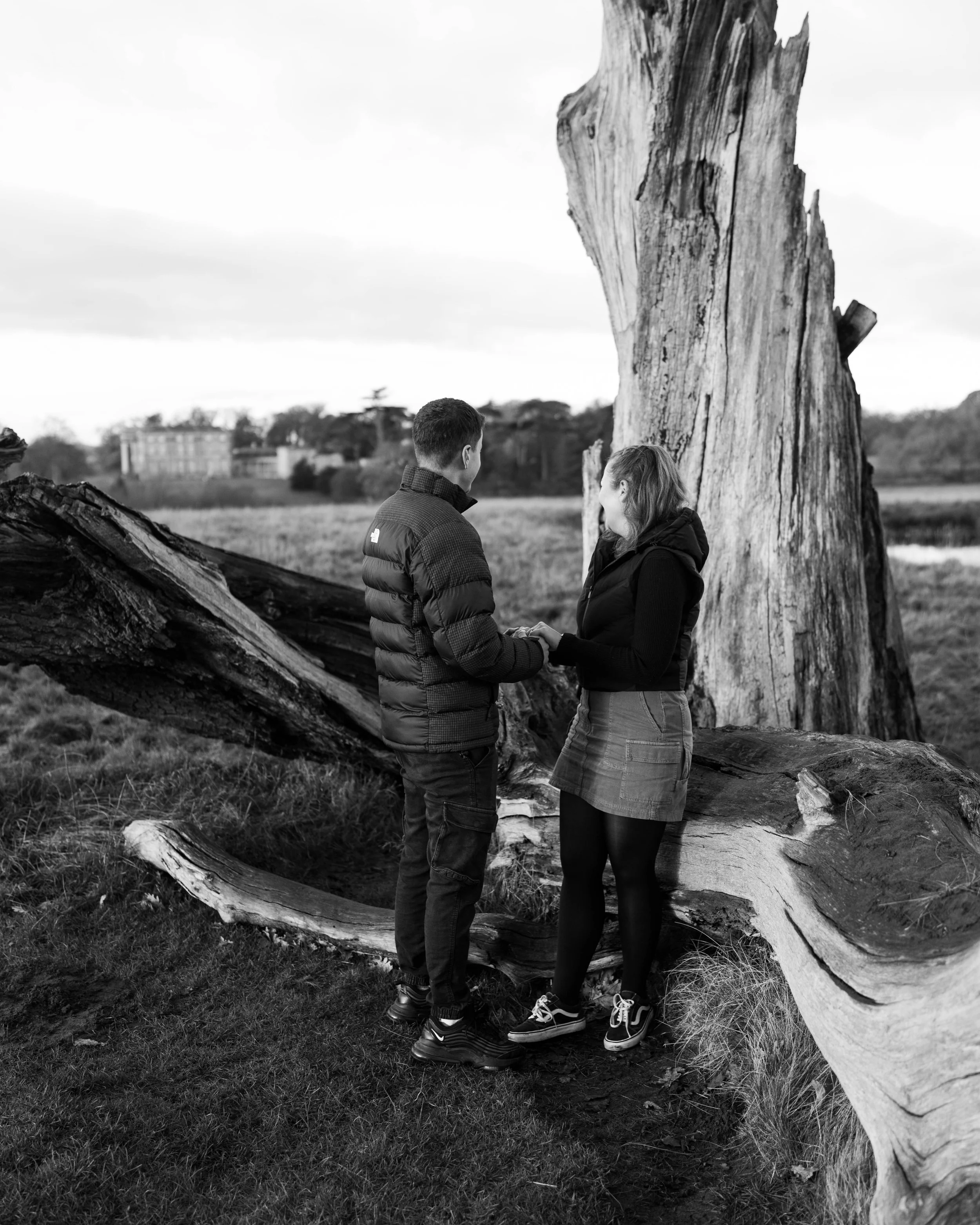 A black-and-white photo of a man and woman standing outdoors, holding hands, with a large fallen tree and a tall standing tree in the background.