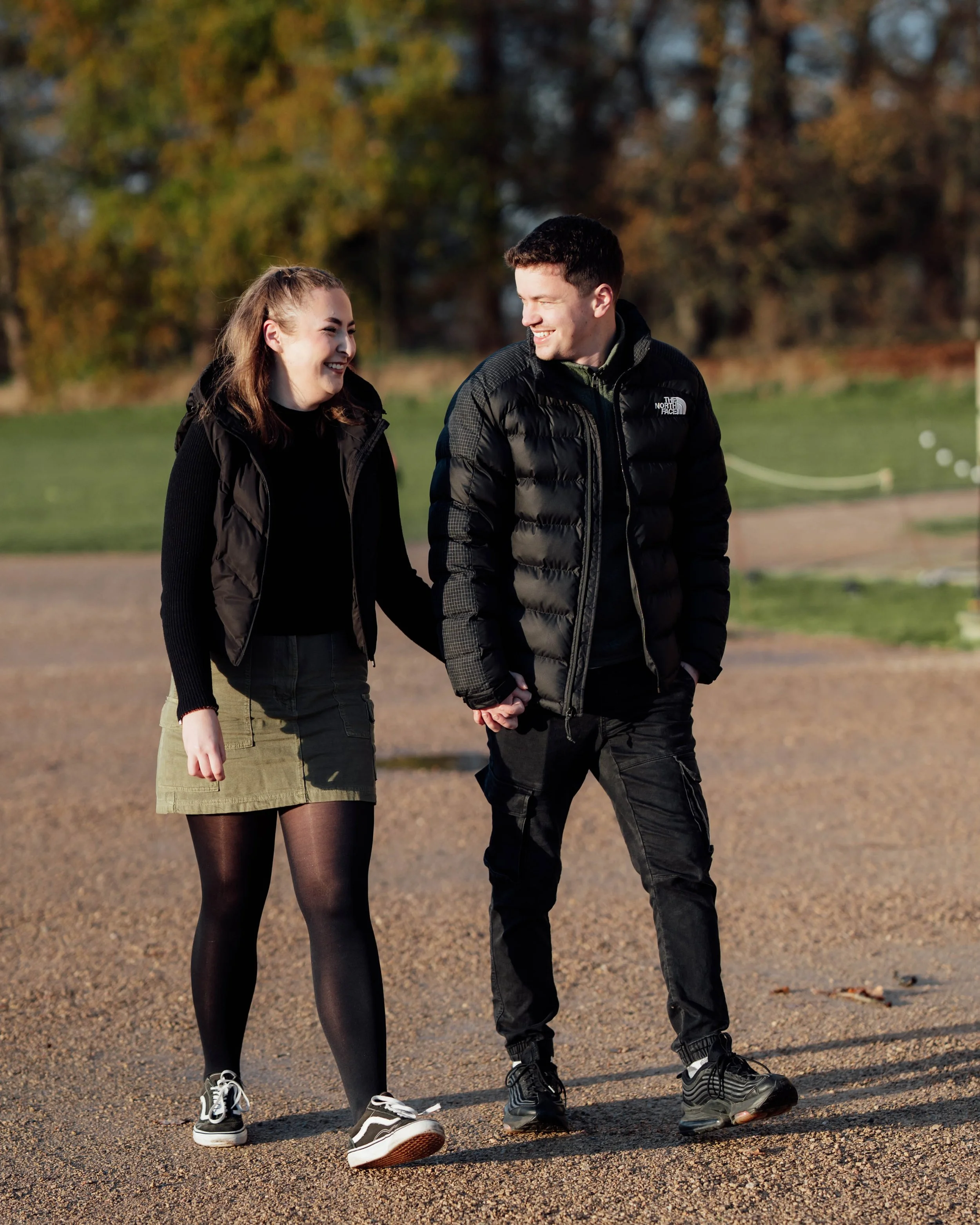 A young couple walking hand in hand outdoors on a gravel path, smiling and enjoying each other's company, with autumn trees in the background.
