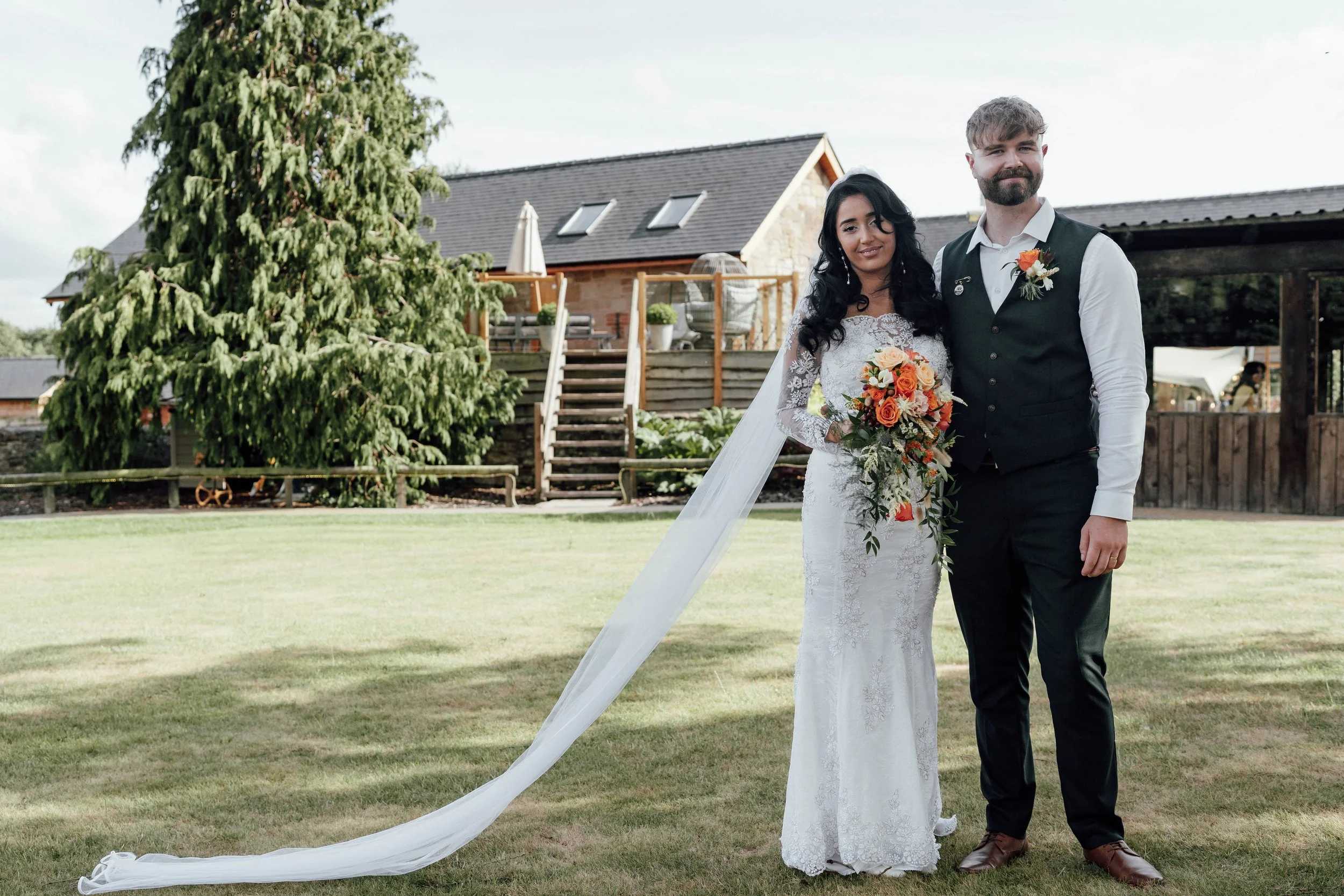 A bride and groom posing outdoors on a sunny day. The bride is wearing a long lace wedding dress and holding a bouquet of orange and cream flowers. The groom is dressed in a white shirt, black vest, and black pants, with a boutonniere matching the br