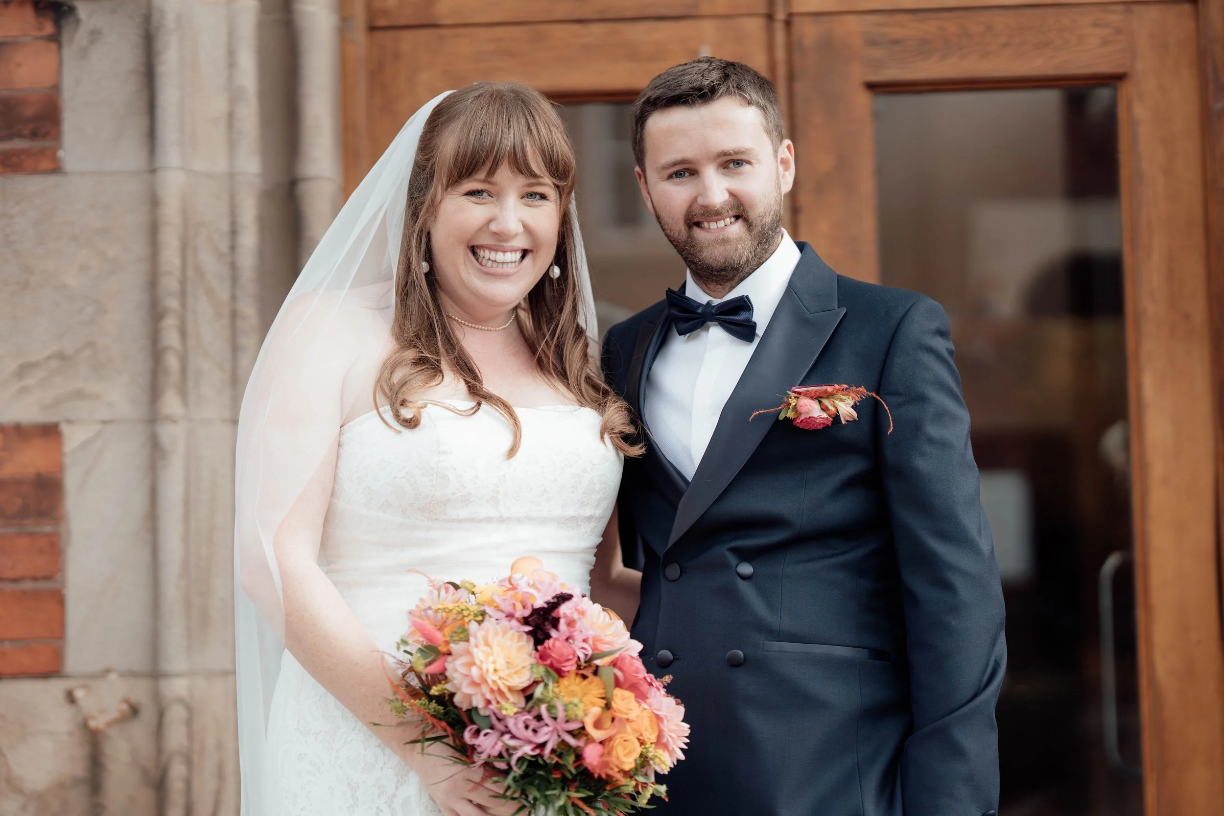 A happy bride and groom standing outside in front of a wooden door, smiling at the camera. The bride wears a white wedding dress and veil, holding a colorful bouquet. The groom wears a dark suit with a bow tie and a boutonniere.