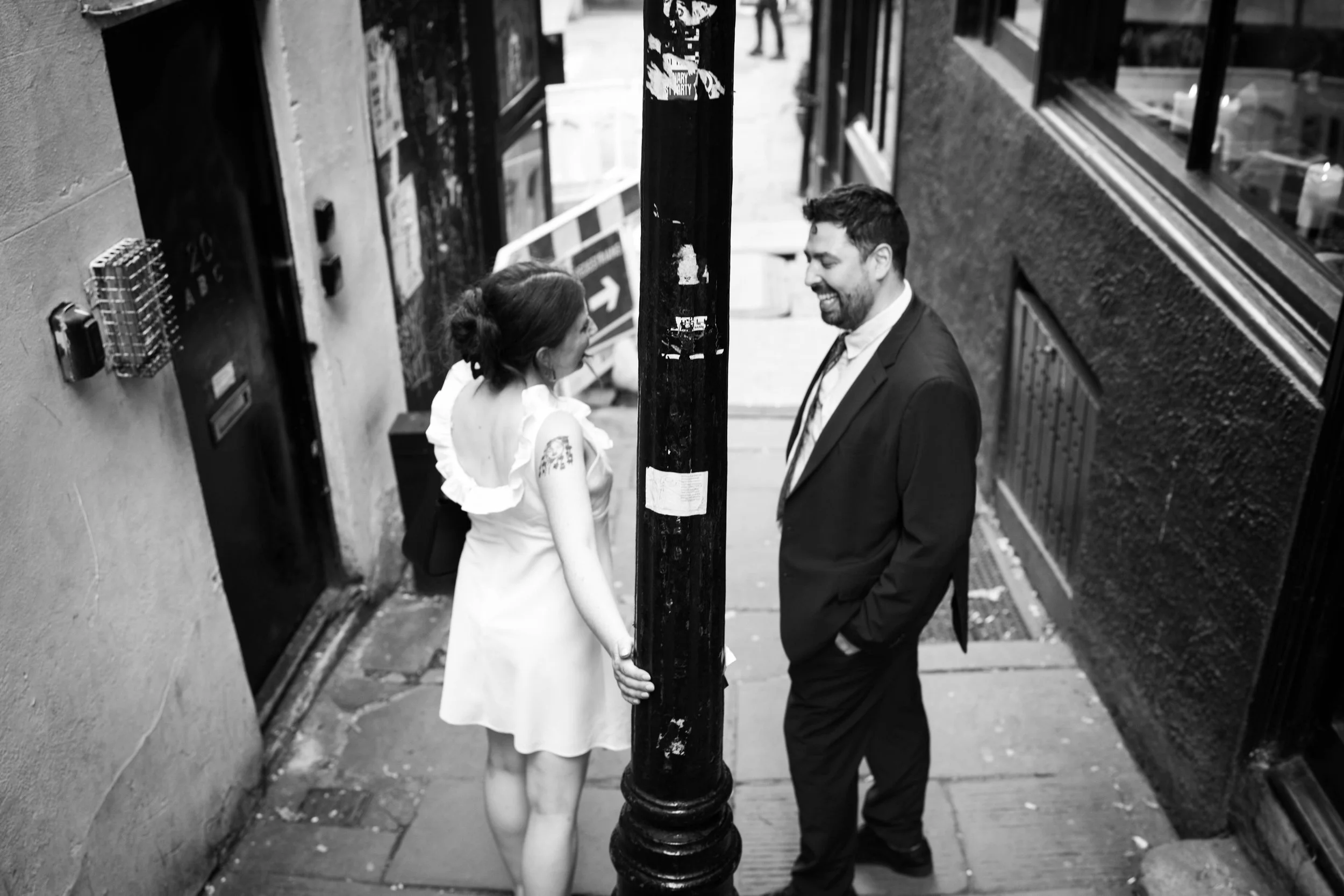 A woman and a man are standing on the sidewalk, chatting and smiling. The woman is holding onto a street pole, with one hand. The woman is wearing a light-colored dress with ruffled sleeves, and the man is dressed in a suit. They appear to be engaged
