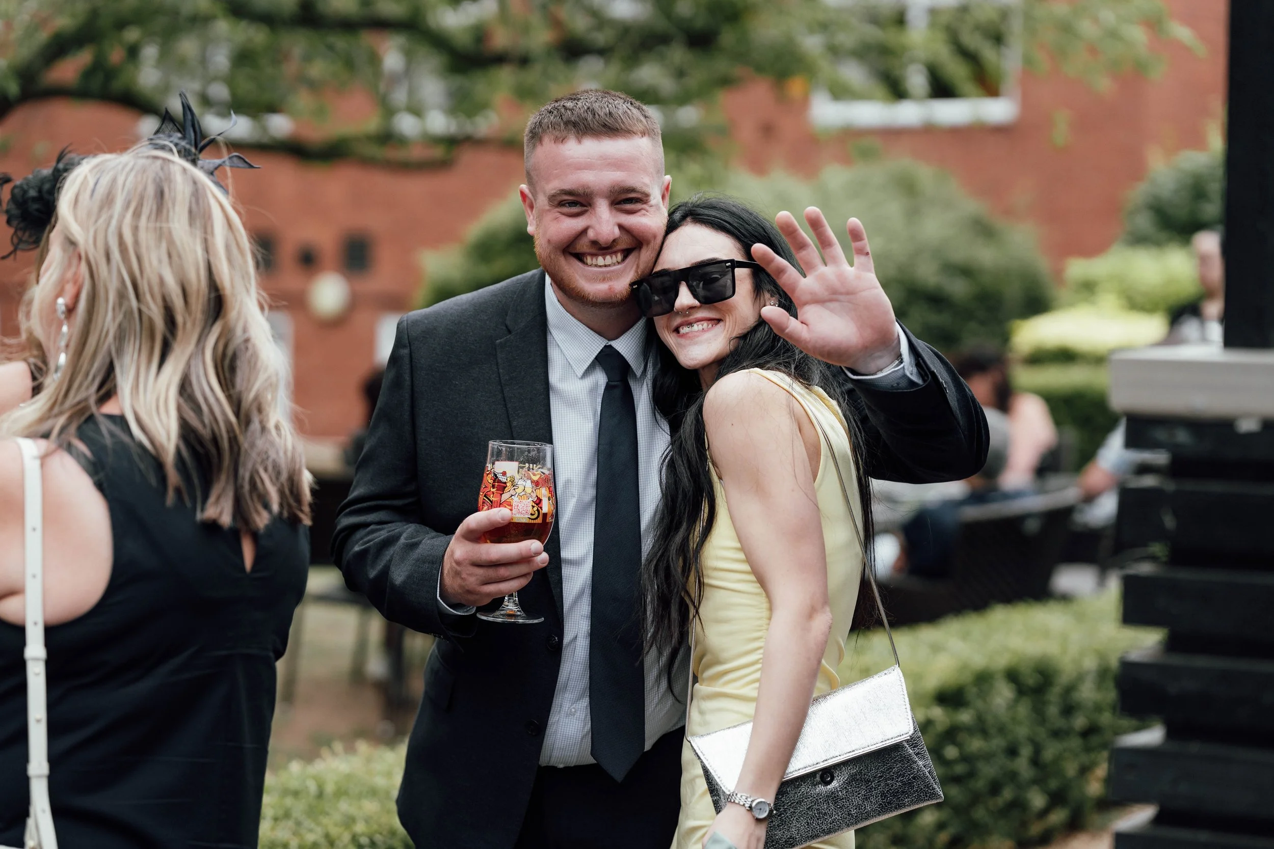A smiling man in a suit holding a drink, standing next to a woman with long black hair, wearing sunglasses and a yellow dress, waving at the camera during an outdoor event.
