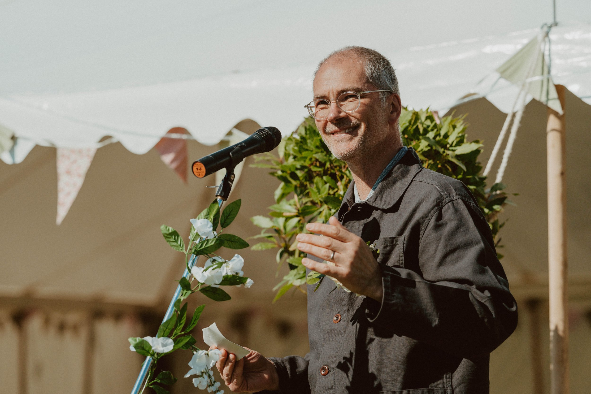 A man with glasses and a gray beard, dressed in a dark jacket, speaking into a microphone at an outdoor event, surrounded by greenery and decorative bunting.