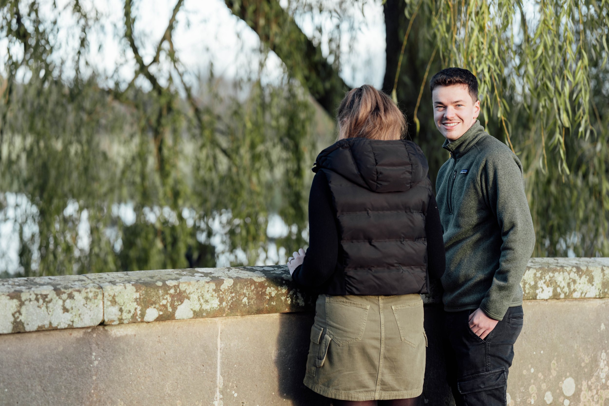A young man and woman standing outdoors by a stone wall, engaged in conversation with trees in the background. The man is smiling and wearing a green zip-up jacket, and the woman has her back to the camera, wearing a black vest and khaki skirt.