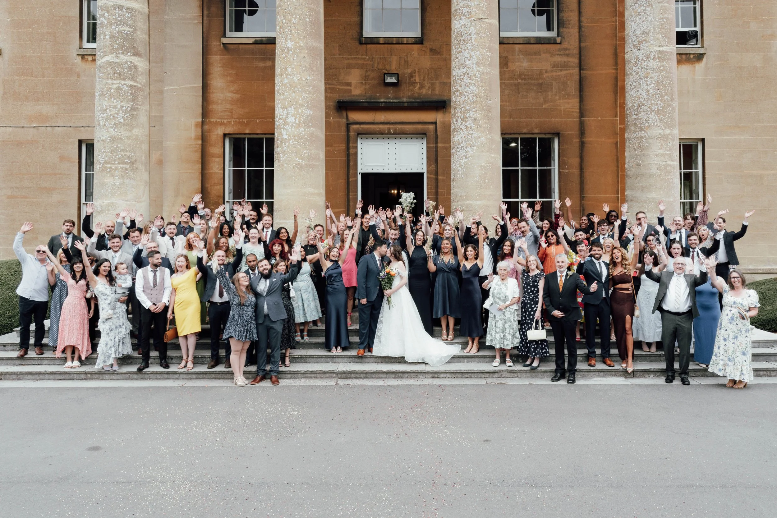 A large group of people celebrating a wedding in front of a historic building with tall columns, smiling and raising their hands in celebration.