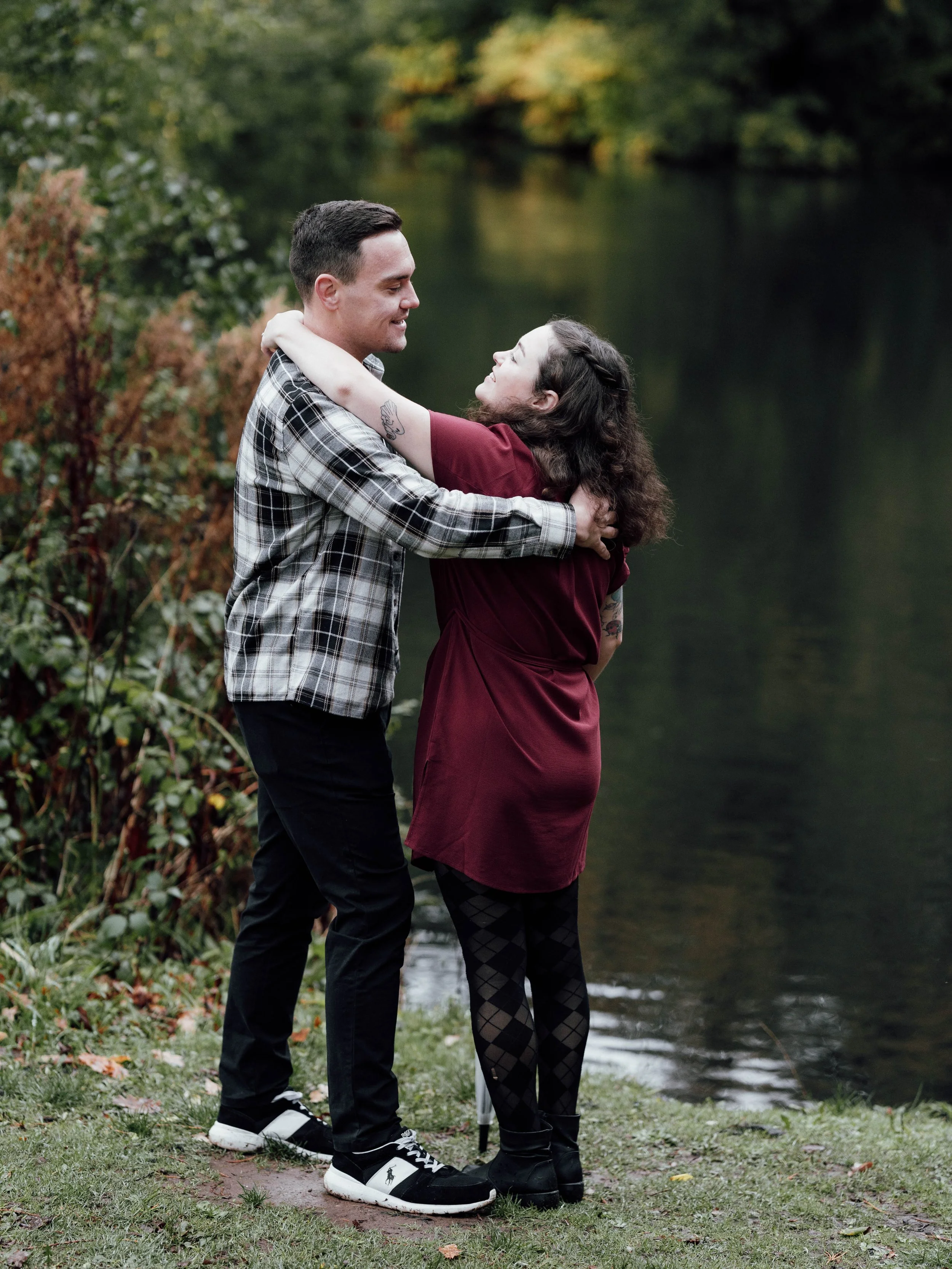 A couple hugging by a lake, surrounded by greenery.