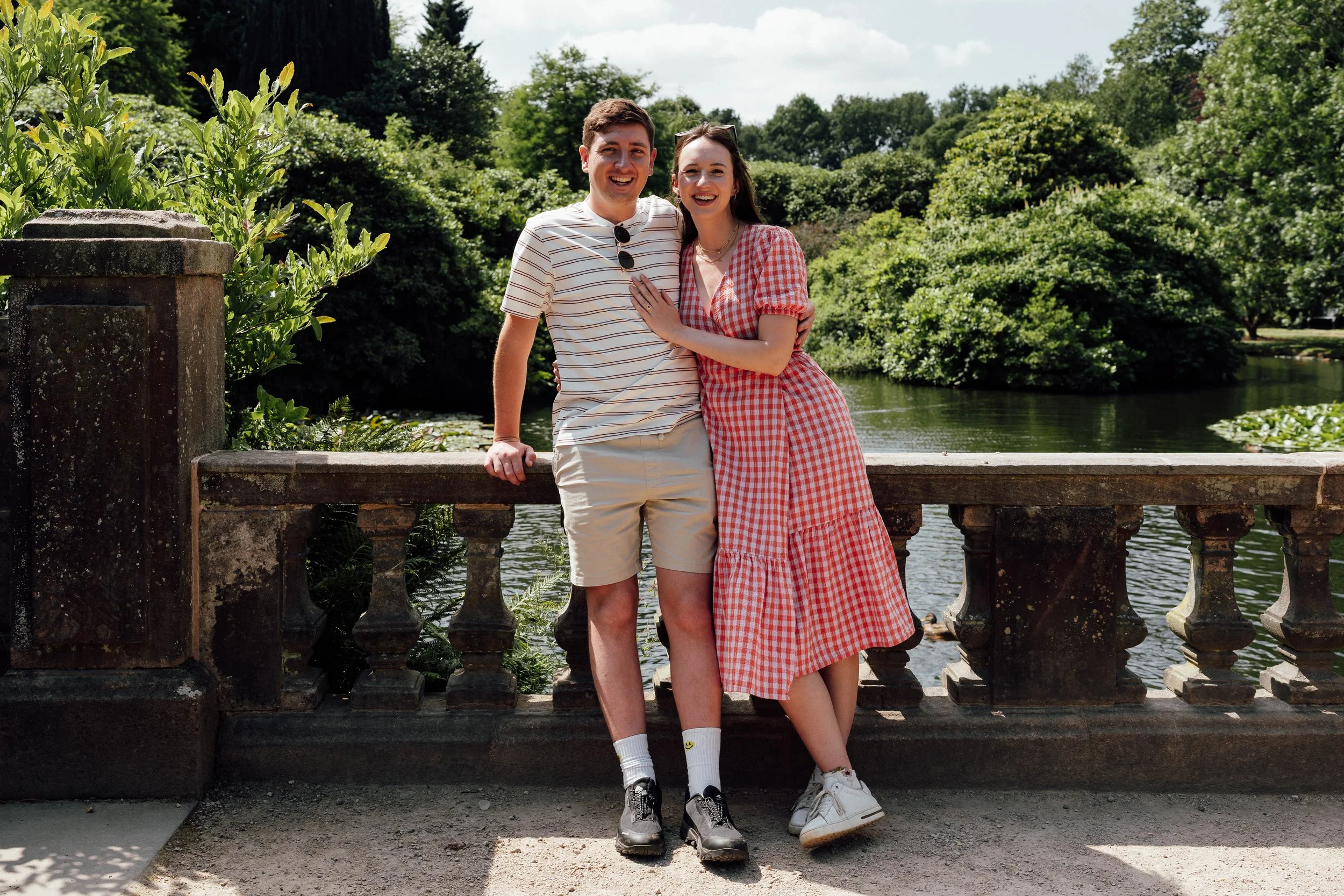 A smiling young man and woman standing close together by a stone railing near a body of water, surrounded by lush green trees and foliage on a sunny day.