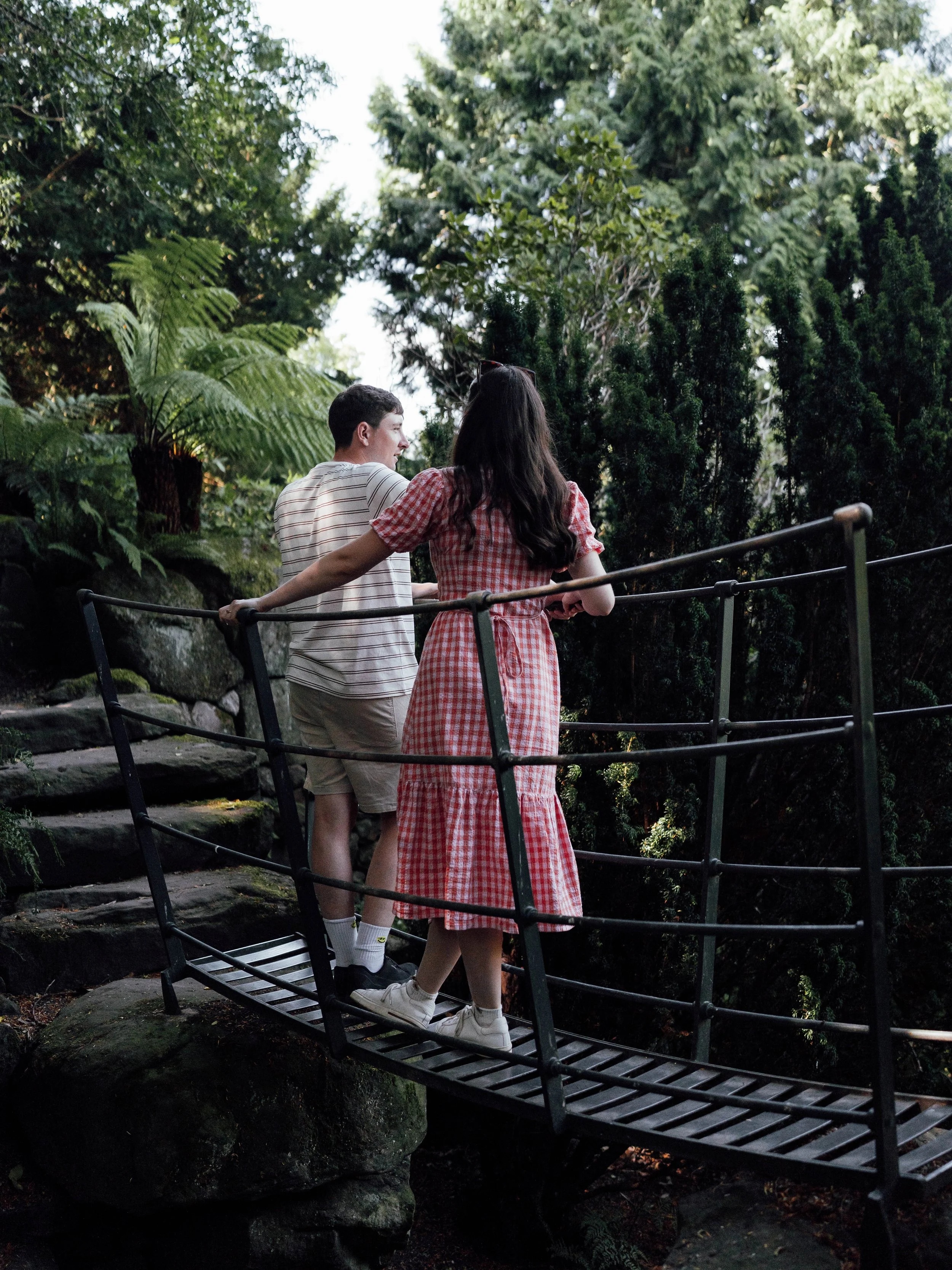 Two people, a man and a woman, standing on a small bridge surrounded by lush greenery in a forest setting, with trees and bushes in the background.
