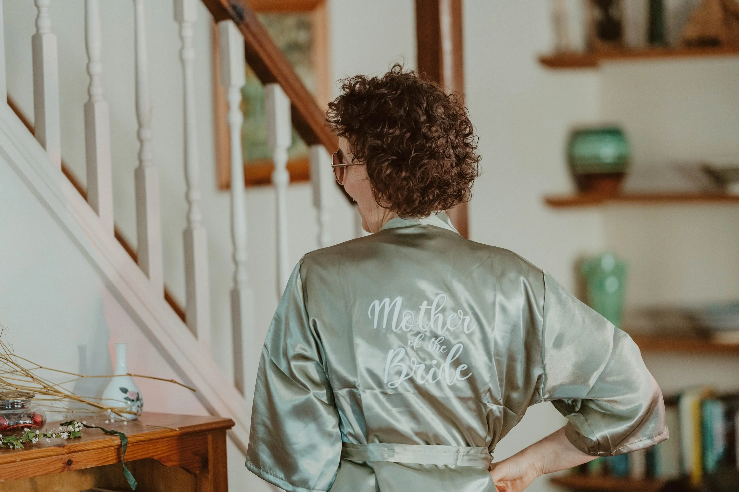 Woman wearing a satin robe with "Mother of the Bride" embroidered on the back, standing in a house near a staircase.