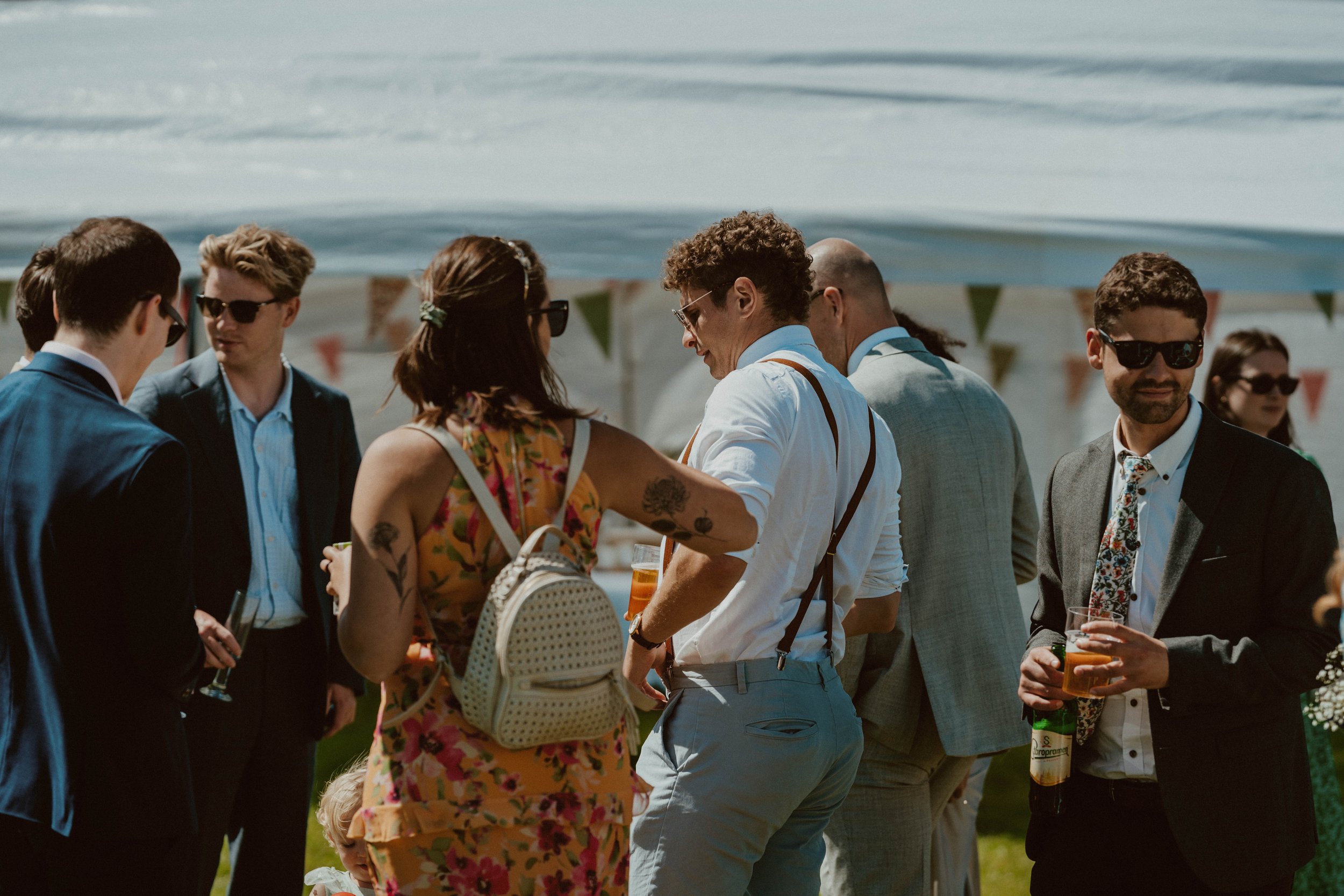 Group of people at an outdoor event by the water, dressed in formal and casual attire, some wearing sunglasses, engaging in conversation, with a white tent decorated with bunting in the background.