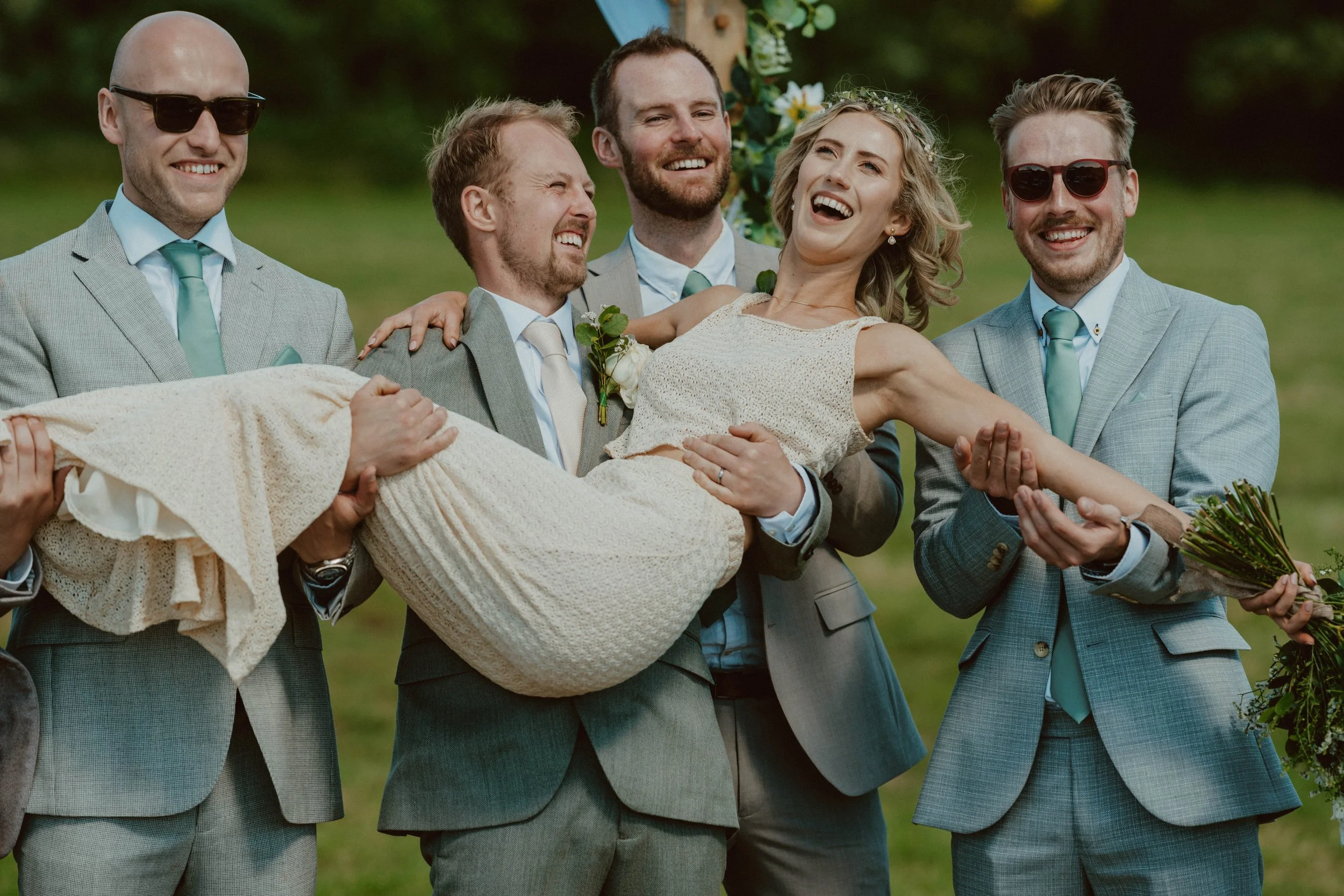 Groom in gray suit and groomsmen in matching light gray suits carrying bride in cream dress during outdoor wedding ceremony.