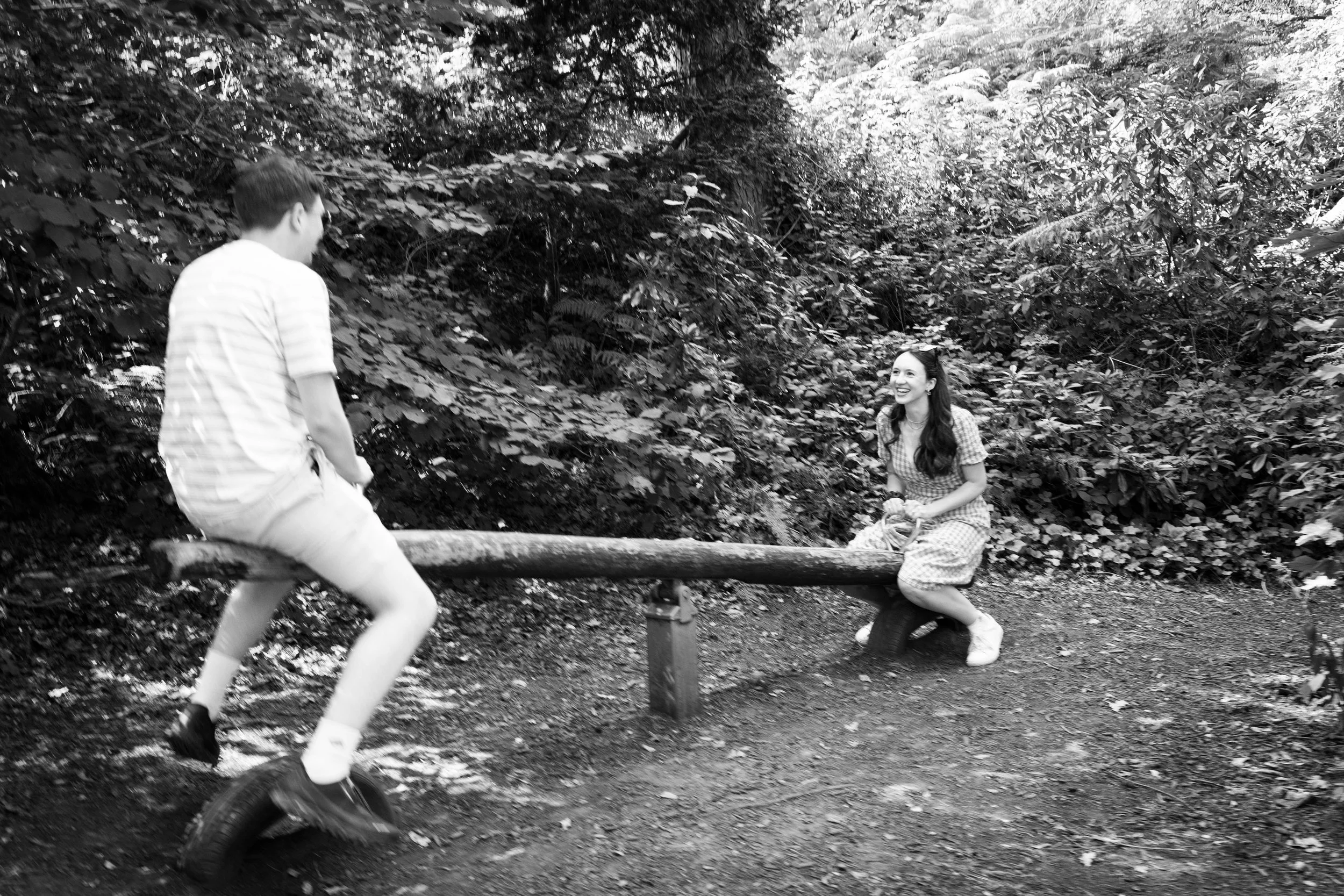 A young man and woman jokingly play on a seesaw in a wooded outdoor area.