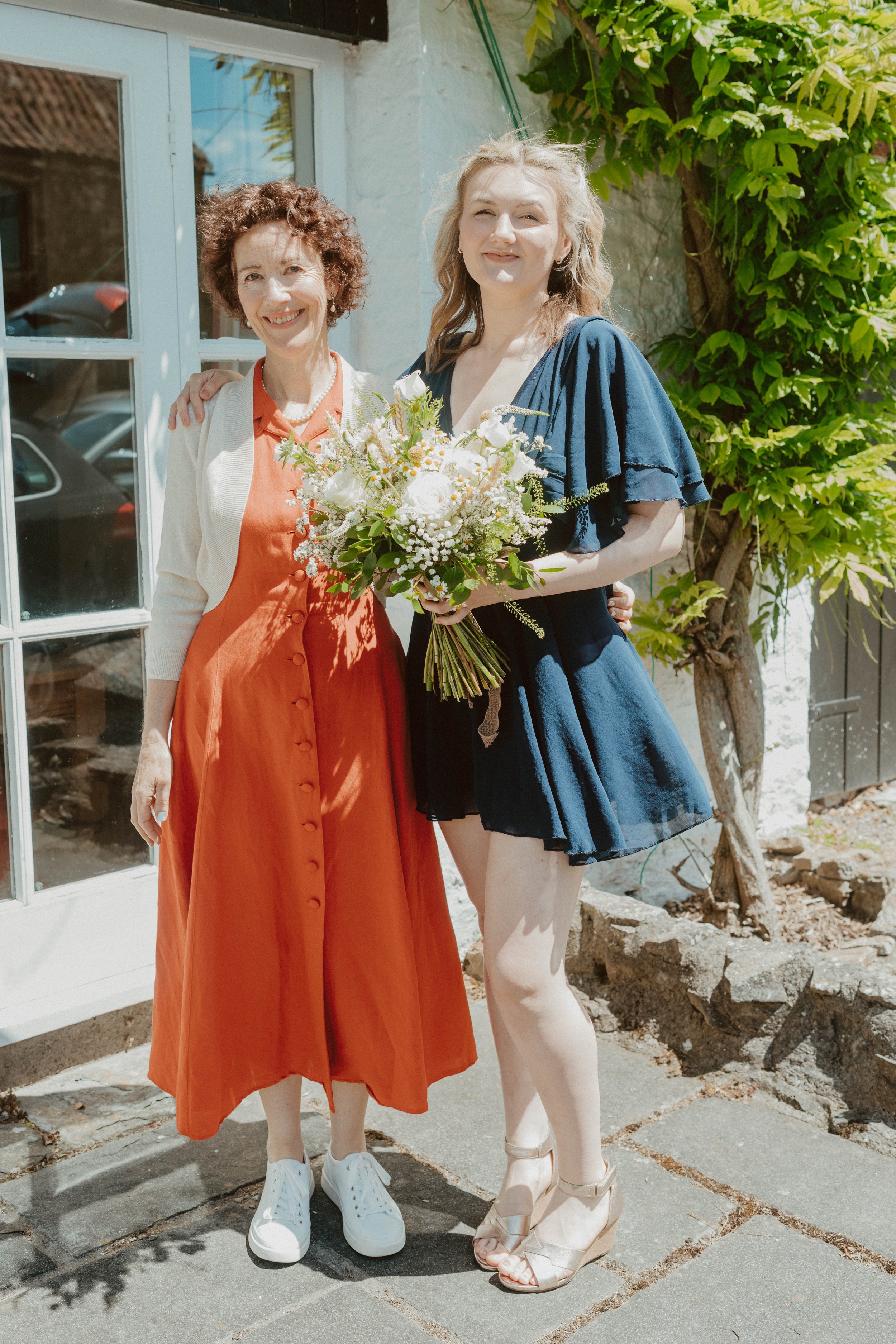 Two women standing outside near a glass door with a reflection of parked cars and a tree, one holding a bouquet of white flowers, both smiling. One woman is wearing an orange dress with white sneakers, and the other in a navy dress with high heels. T