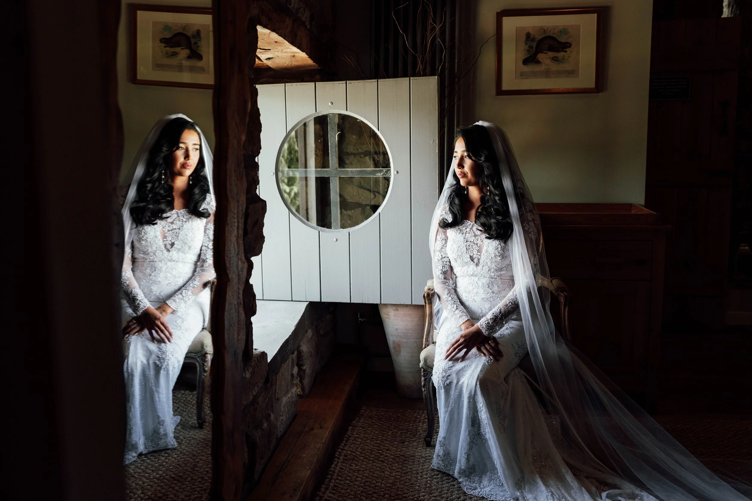 A bride in a white lace wedding dress and veil sitting on a vintage chair in front of a mirror, with her reflection visible. The room has wooden walls and framed pictures, with sunlight coming through a circular window, creating a soft ambiance.
